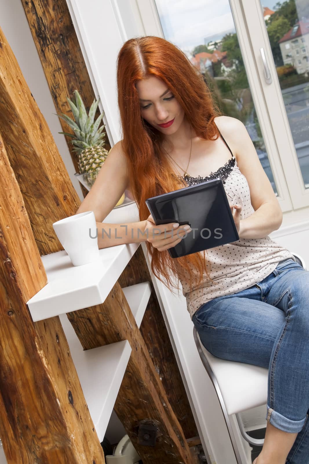 Pretty Young Blond Woman Sitting Near the Window with a Glass of Drink and a Tablet Computer.