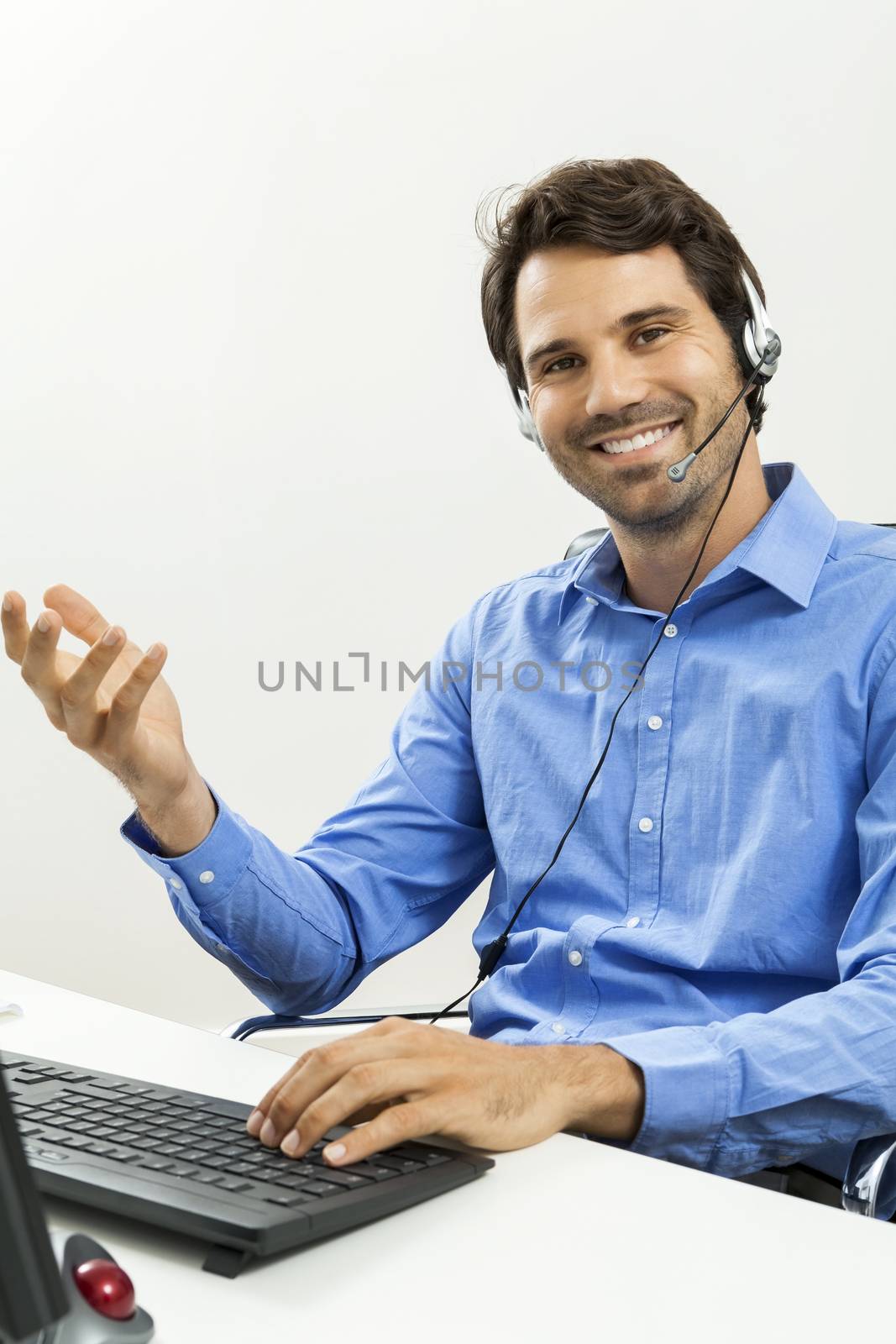 Attractive unshaven young man wearing a headset offering online chat and support on a client services of help desk as he types in information on his computer