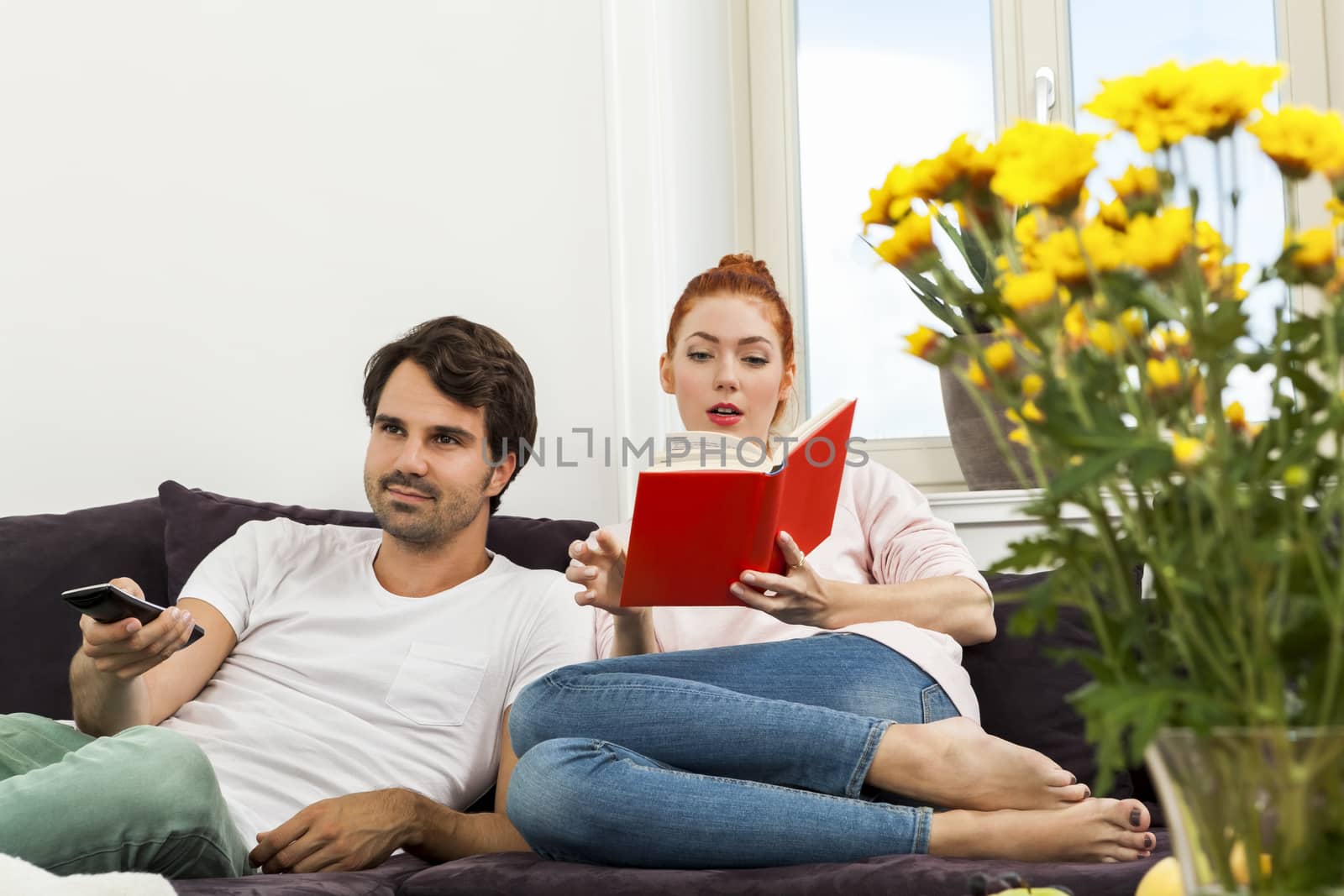 Young Couple Resting on the Sofa at the Living Room While the Man is Watching TV and Woman is Reading a Book.