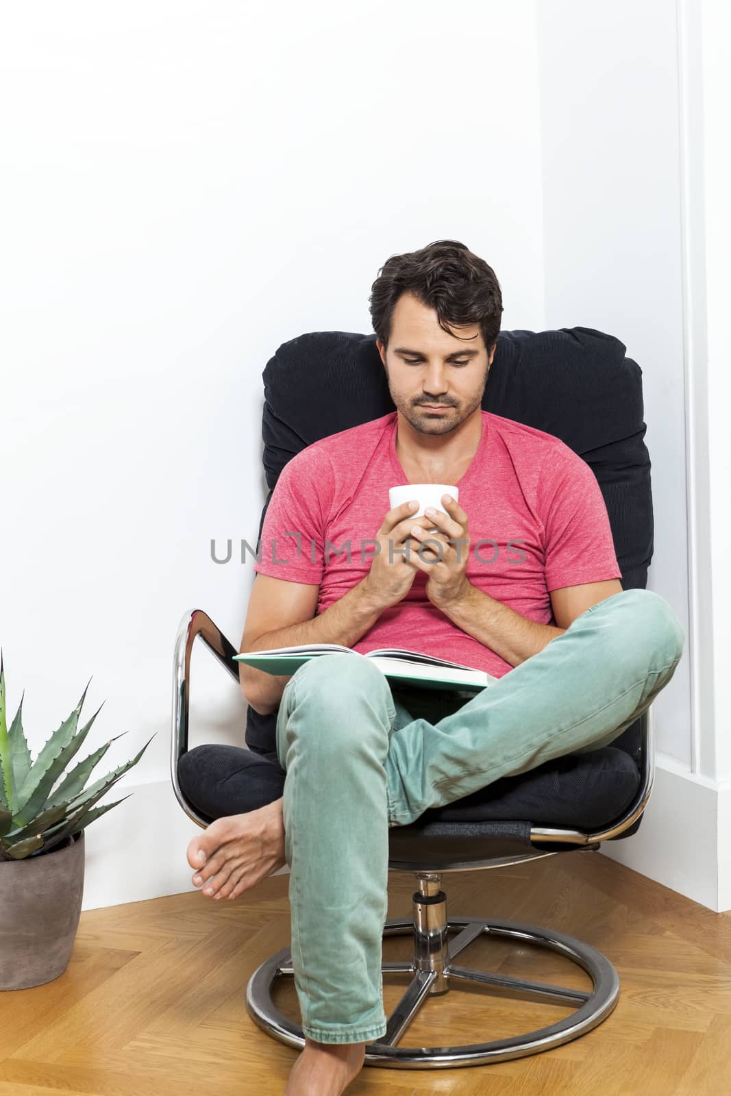 Young Man in Casual Clothing Sitting on Black Chair While Reading a Book and Holding a Glass of Drink.