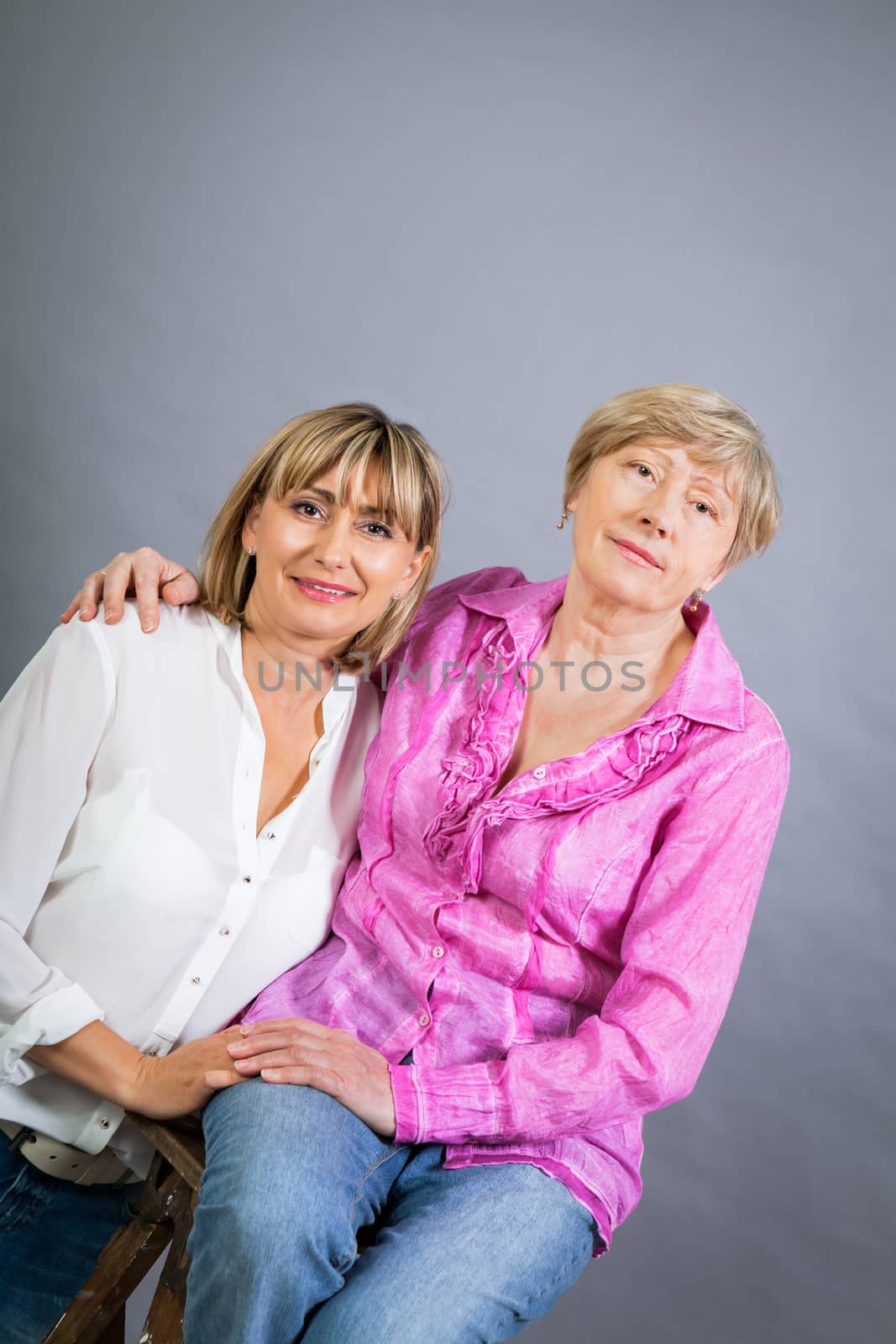 Attractive stylish blond senior lady with her beautiful middle-aged daughter posing together with her hands on her shoulders smiling at the camera on a grey studio background