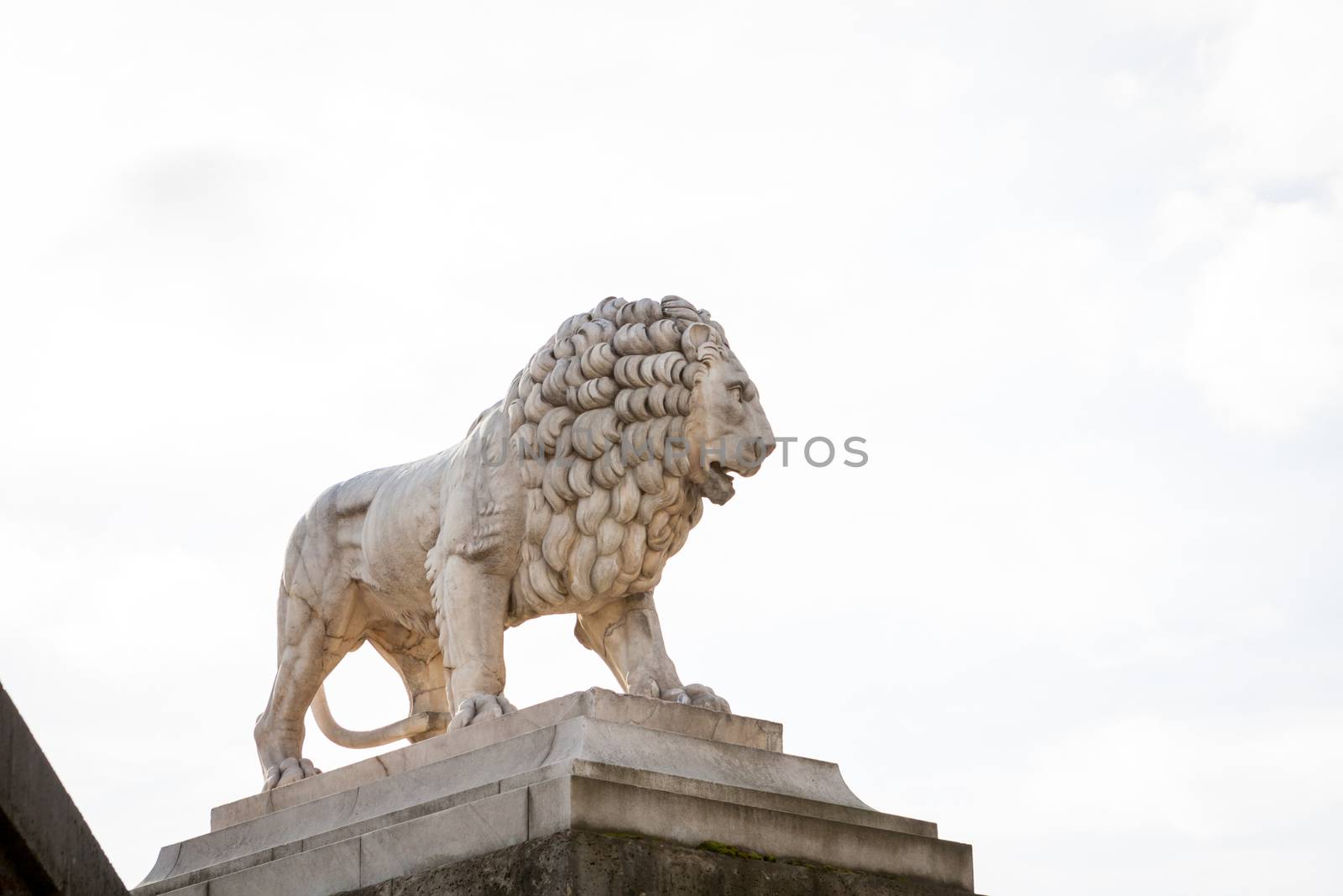 Bird perched on an ancient stone statue of a naked seated god or deity with a long curly beard and hair in Paris, France, possibly part of a stone fountain, viewed low angle against a cloudy blue sky