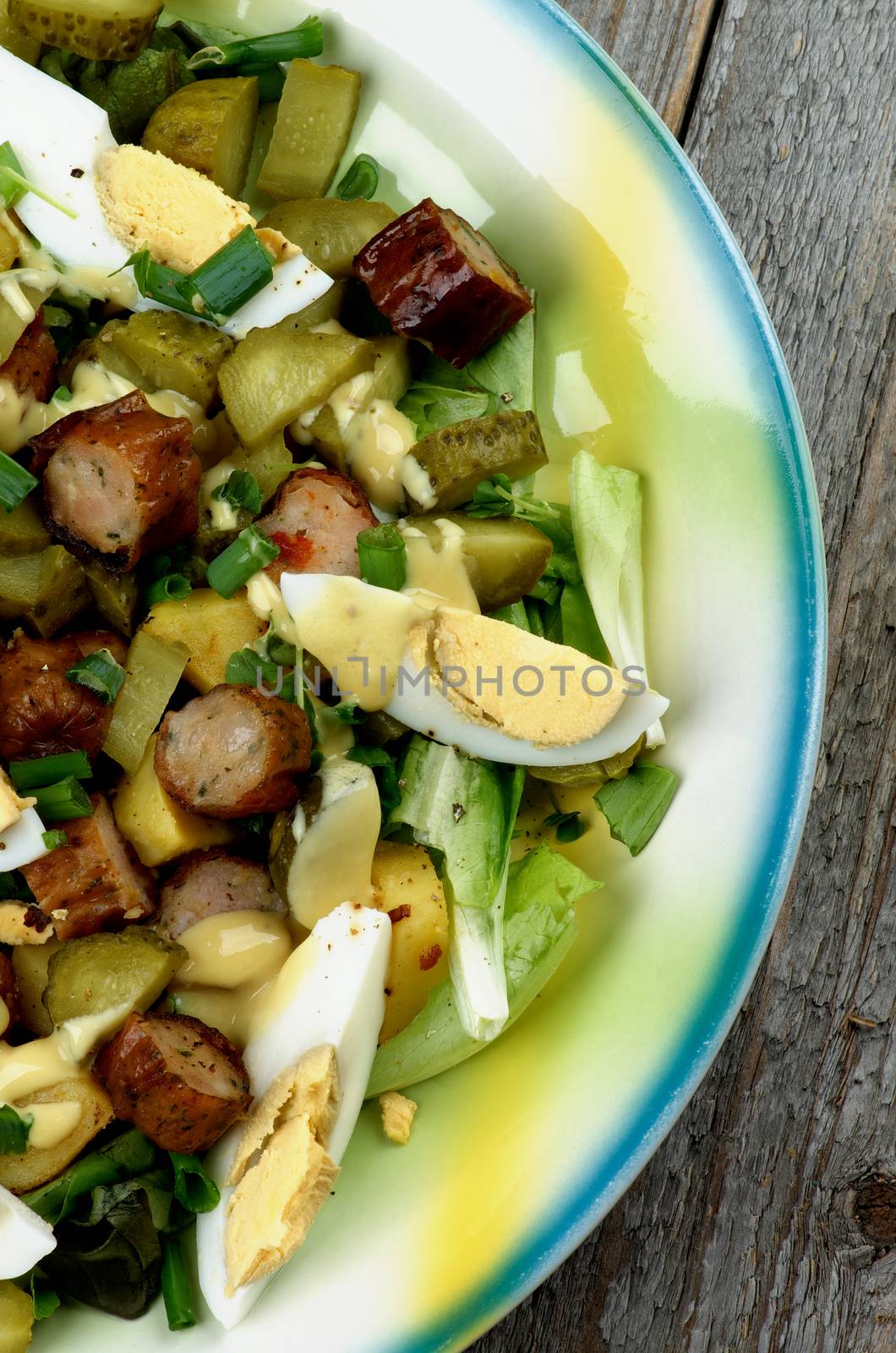 Delicious Potato and Sausage Salad with Gherkins, Lettuce, Boiled Eggs and Mustard Sauce closeup on Yellow and Blue Plate closeup on Rustic Wooden background