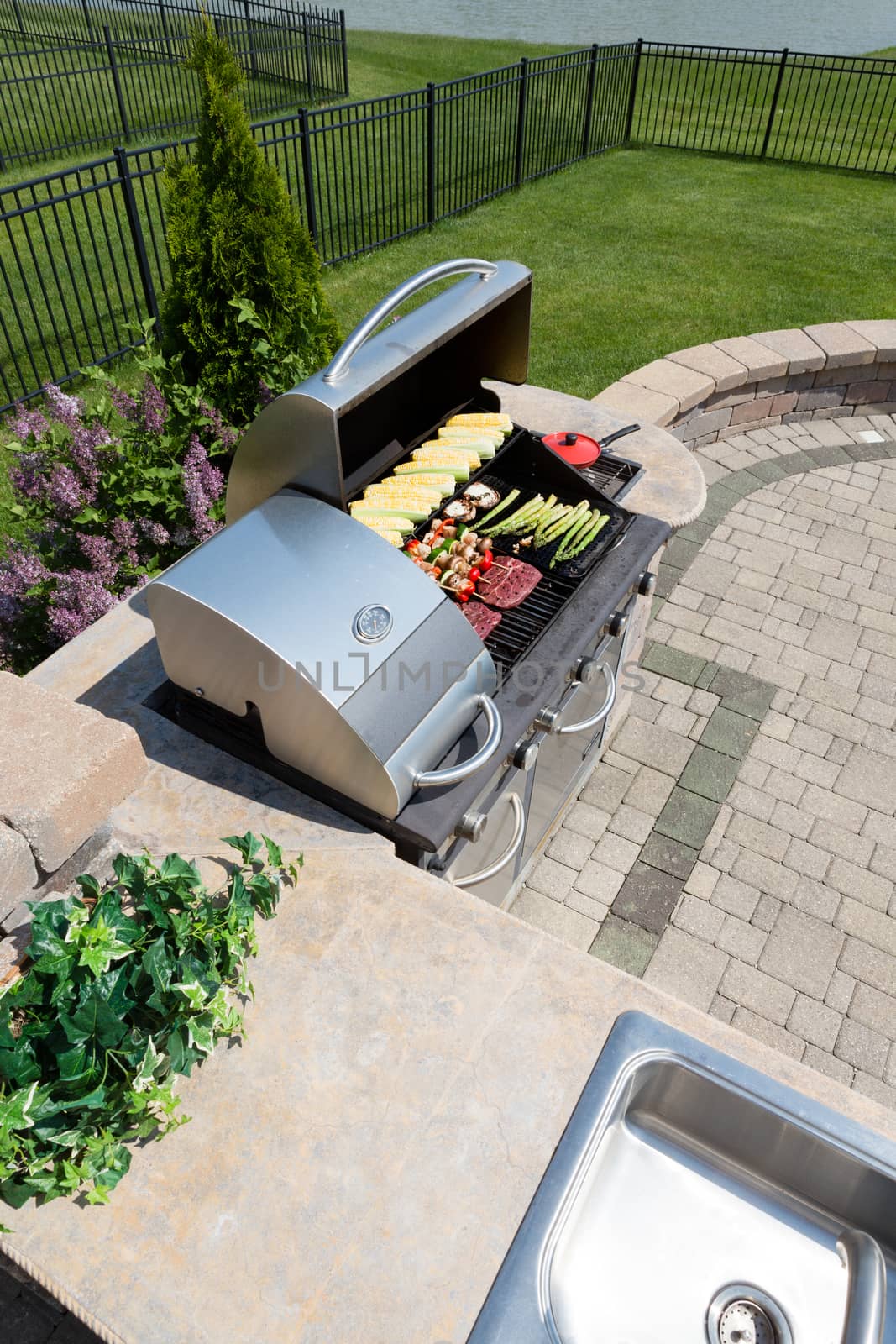 Healthy outdoor living cooking in a summer kitchen fitted with a sink and counter and large gas barbecue loaded with fresh vegetables and meat on an open-air brick patio in the back garden