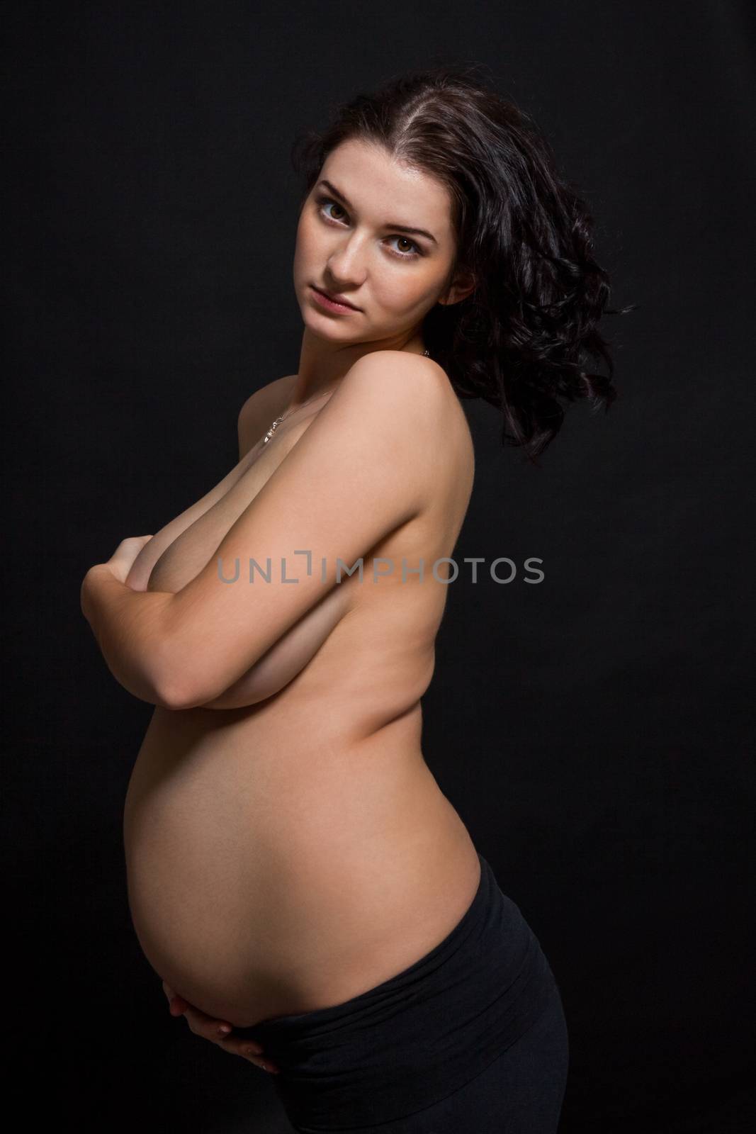 Pregnant woman posing nude cupping her large swollen abdomen tenderly with her hand while covering her breasts with the other, standing sideways on a dark background smiling at the camera