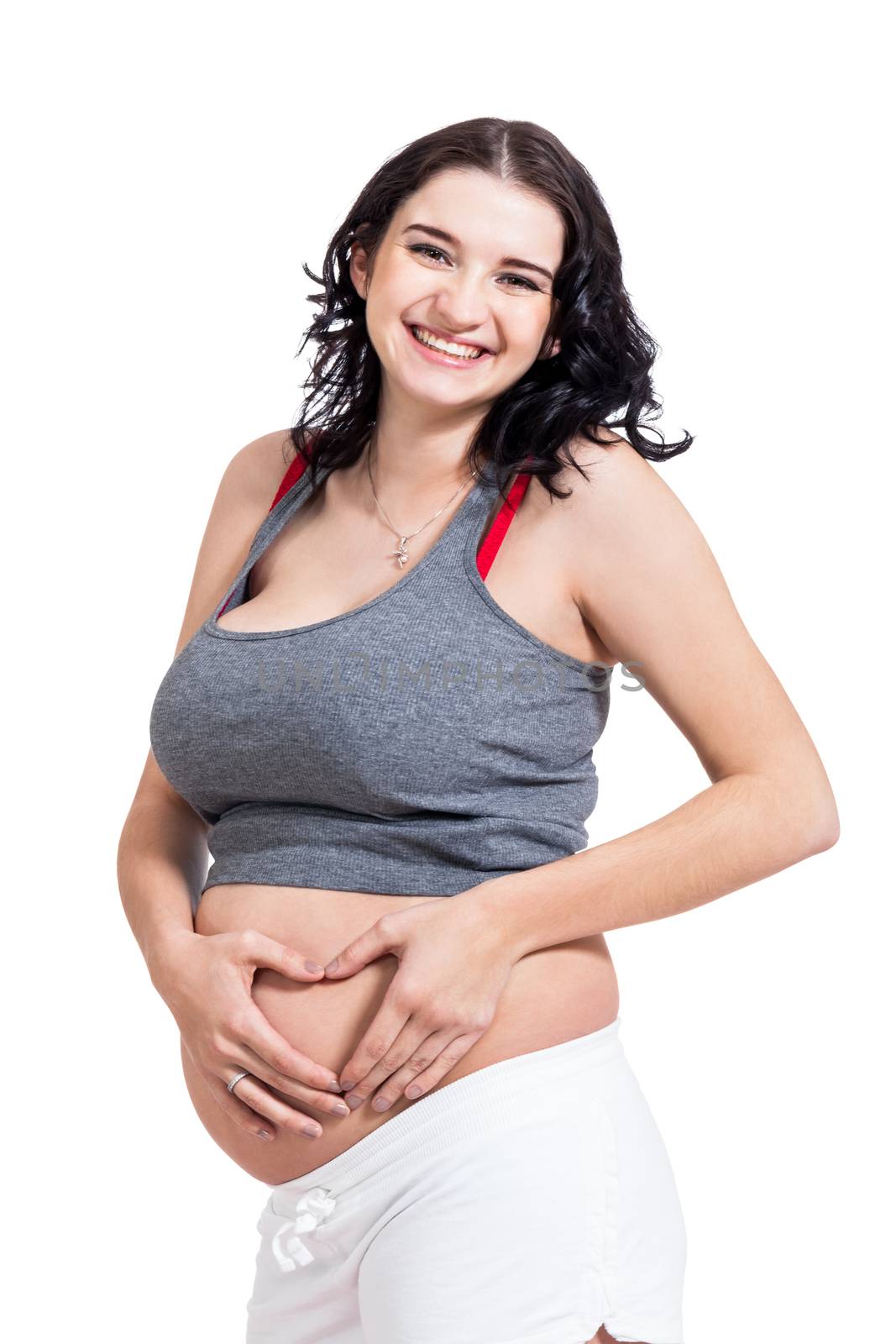 Young pregnant woman making a heart gesture with her hands against her bare belly showing her love of her unborn child while looking at the camera with a smile, isolated on white