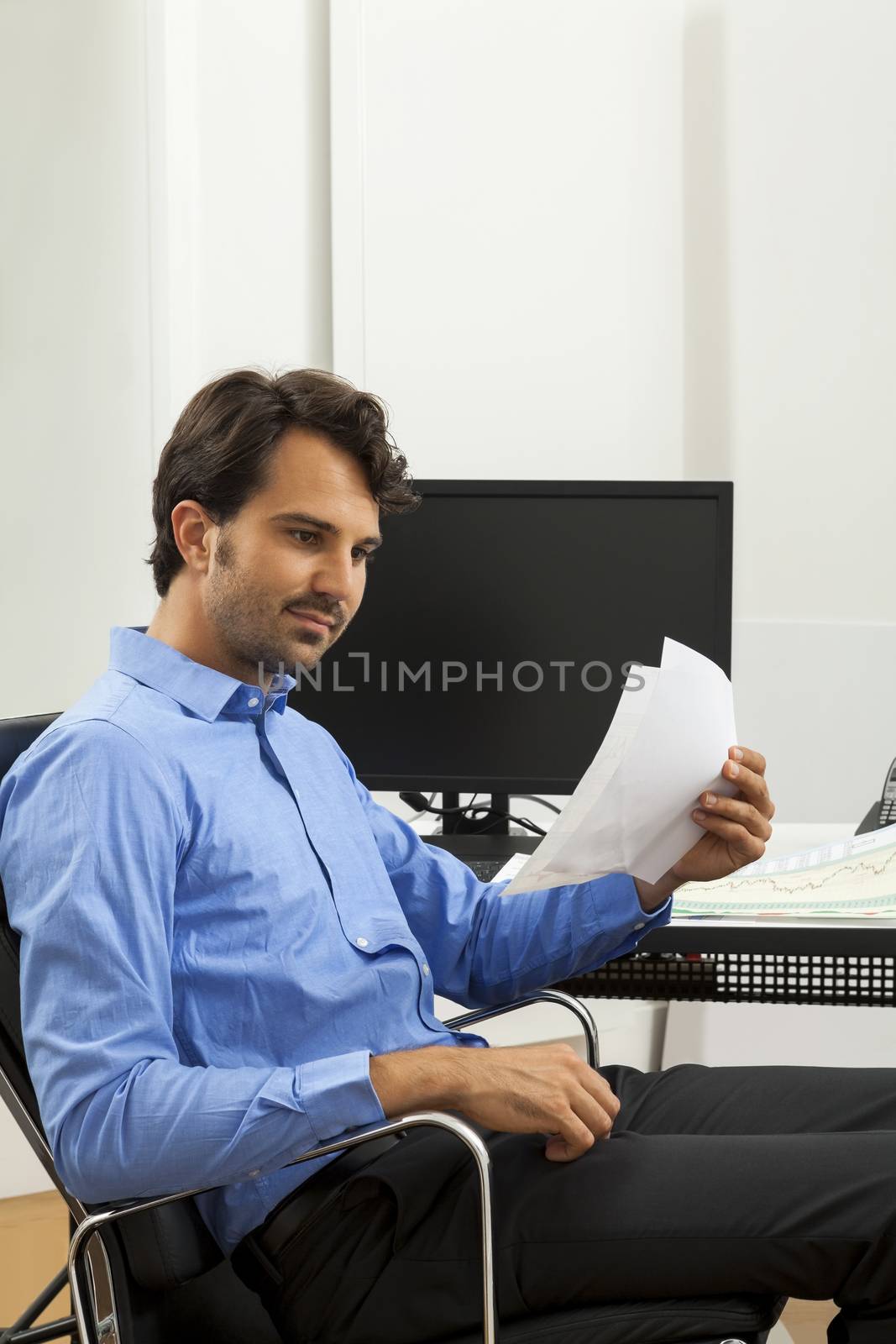 Young handsome man sitting at his desk in the office while reading written agreements and studying important documentation for work