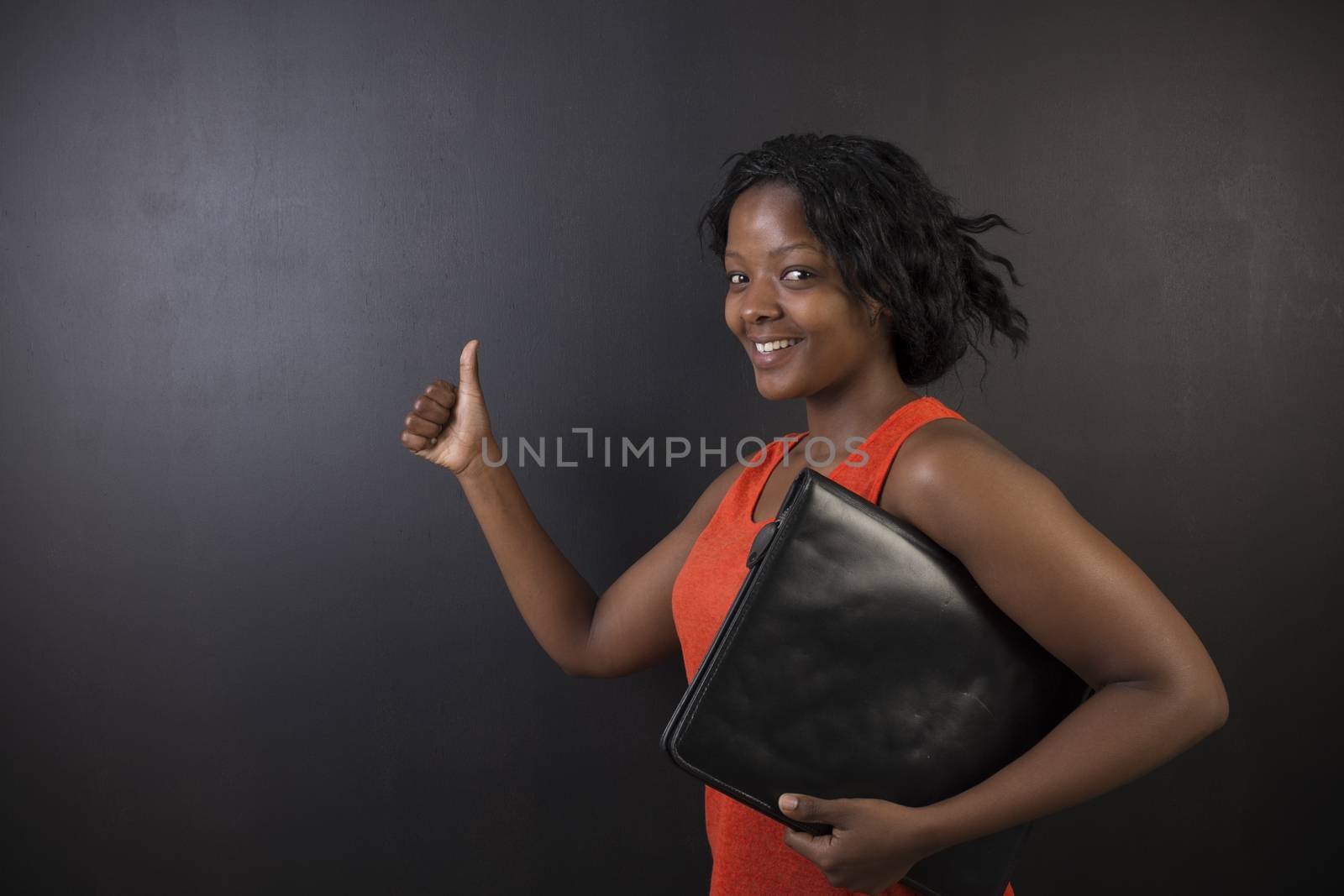 South African or African American woman teacher or student with thumb up holding diary notepad on chalk black board background