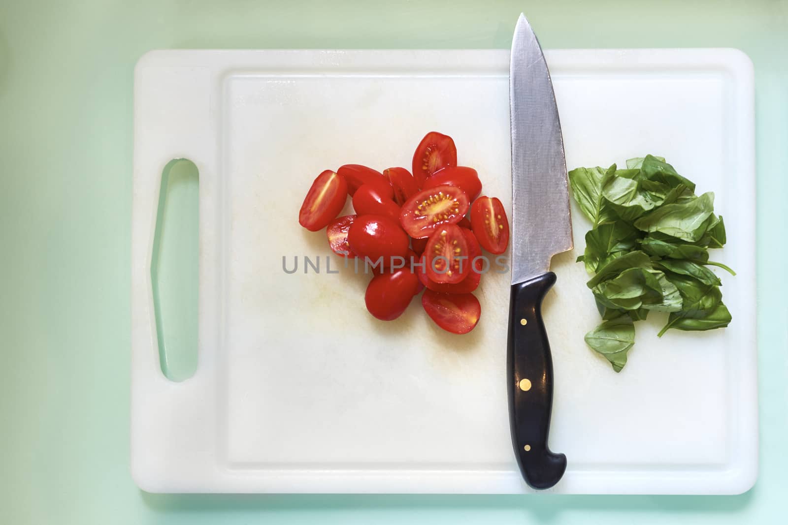 High angle shot of kitchen knife resting on white chopping board with cherry tomatoes in half and fresh basil leaves.