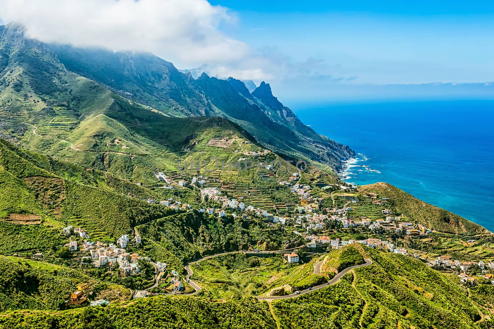 Small city or village buildings and winding or serpantine road in green mountain or rock valley and clouds with horizon landscape near coast of Atlantic ocean in Tenerife Canary island, Spain