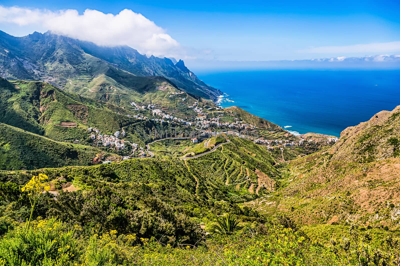 Small village buildings and winding or serpantine road in green mountain or rock valley and clouds with horizon landscape near coast of Atlantic ocean in Tenerife Canary island, Spain