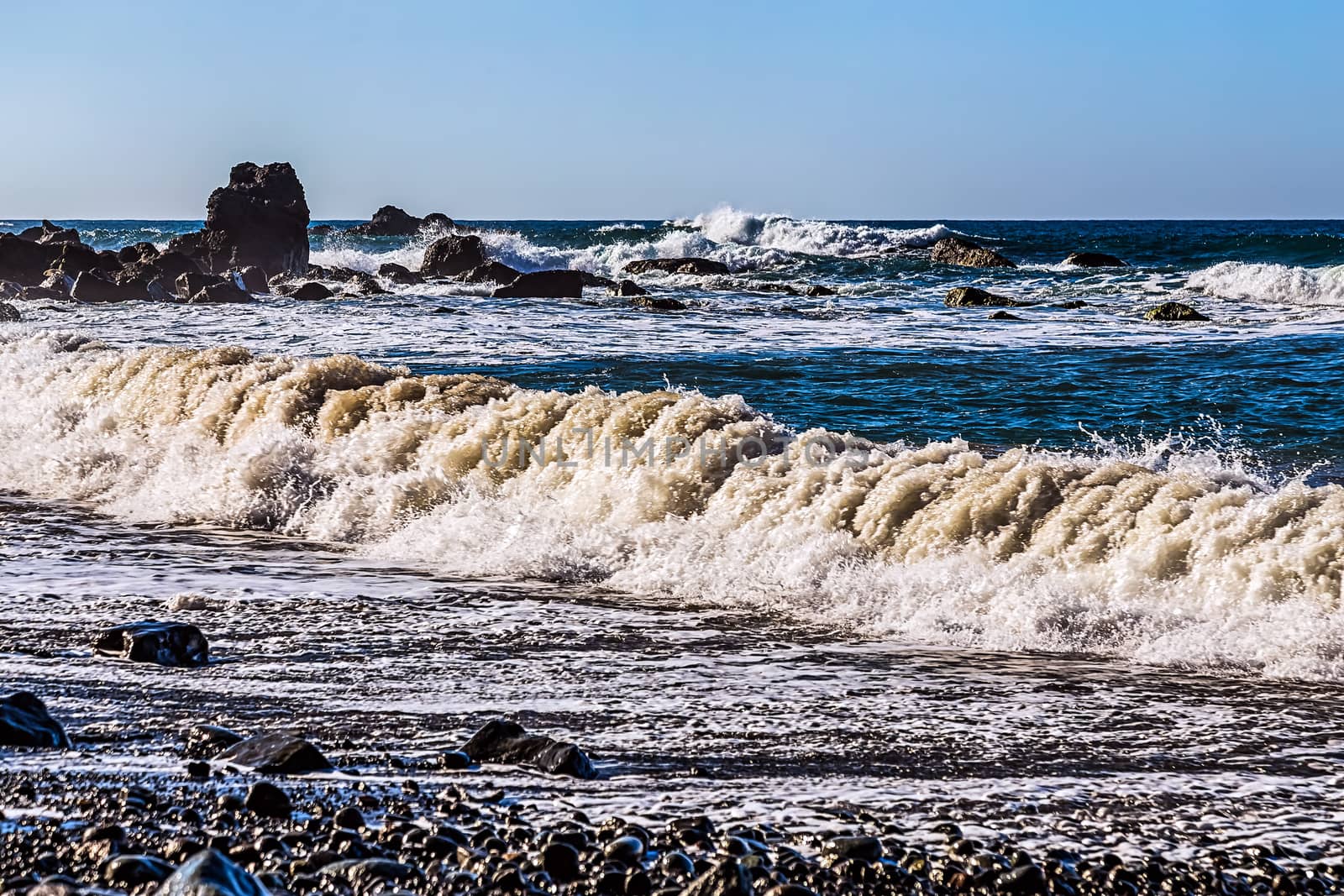 Waves and foam on wild stone beach on coast or shore of the Atlantic ocean with sky and rock on skyline or horizon in Tenerife Canary island, Spain at spring or summer