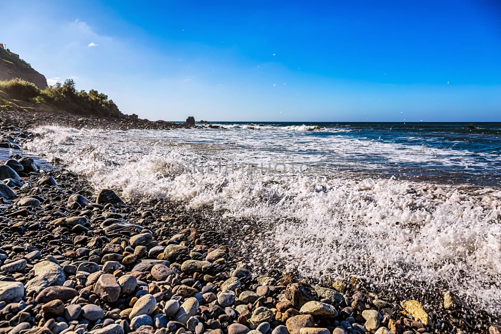 Wild stone beach on coast or shore of the Atlantic ocean with waves and foam and blue sky and  horizon in Tenerife Canary island, Spain