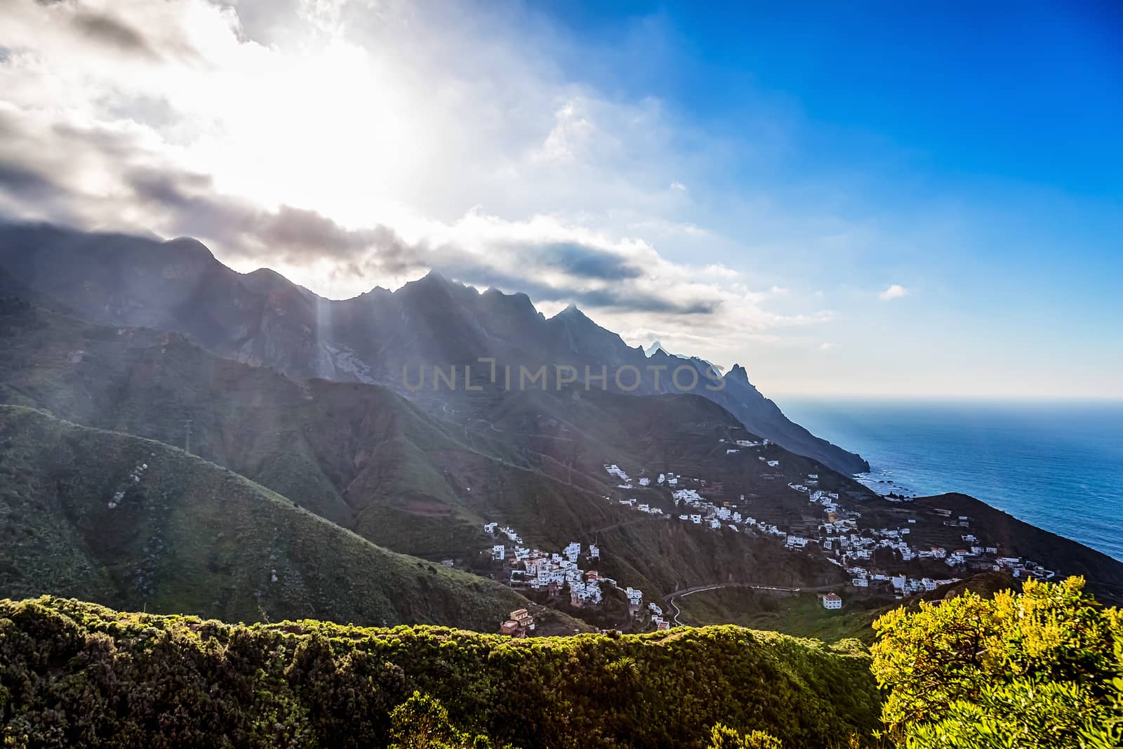 Green mountains or rock with clouds and sun with sunlight and sunbeams on sky over small village or city near the Atlantic ocean in Tenerife Canary island, Spain