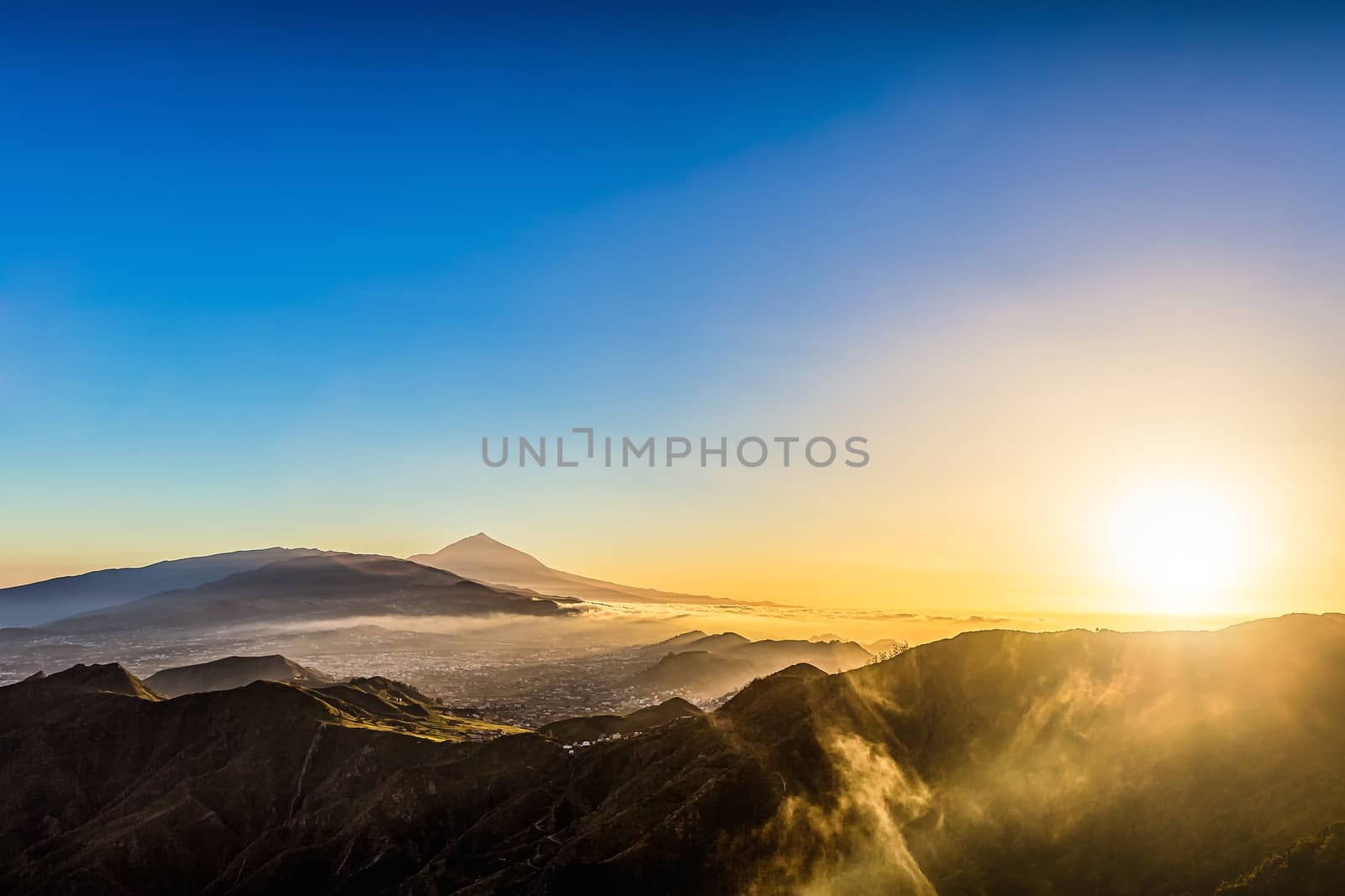 Sun with sunlight over mountains on blue sky and clouds with haze and Teide volcano on background at evening sunset in Tenerife Canary island, Spain at spring or summer
