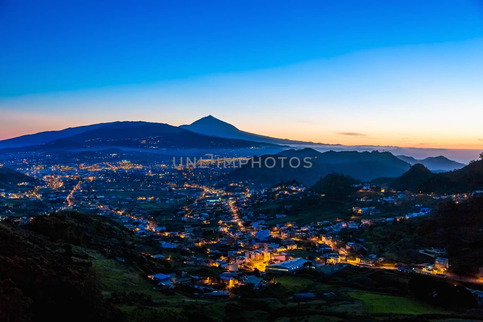 City or town with illumination after sunset at evening in mountains with blue sky and Teide volcano on background in Tenerife Canary island, Spain at spring or summer