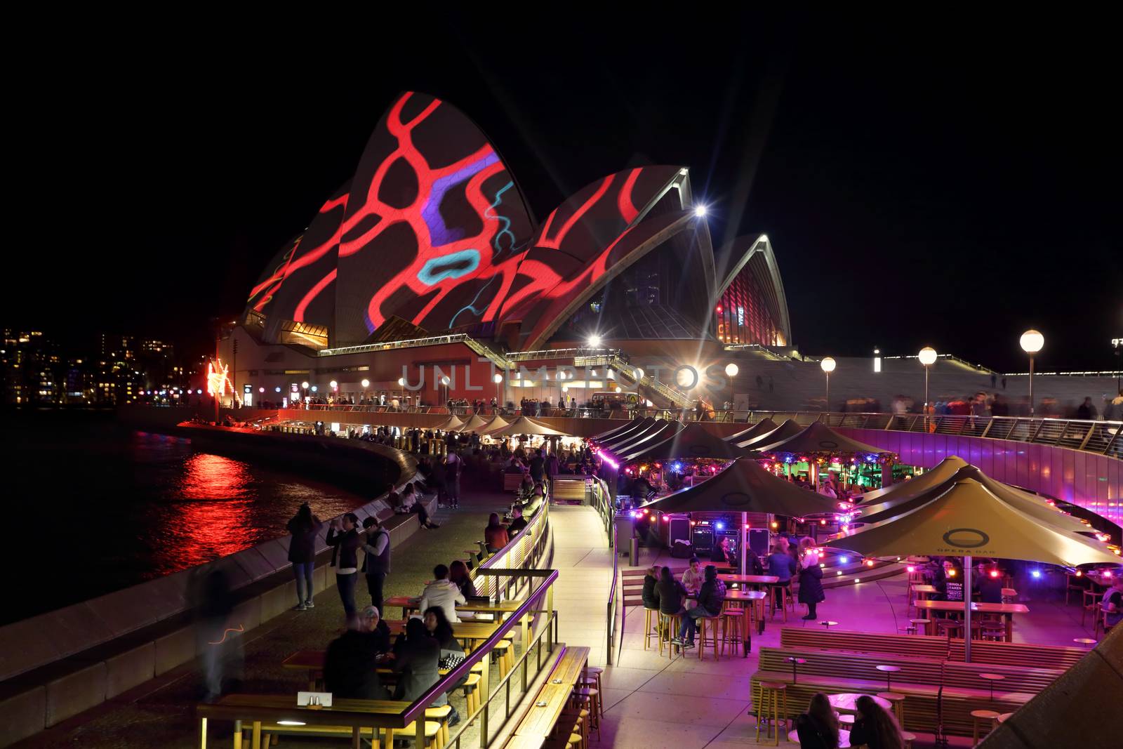 SYDNEY, AUSTRALIA - MAY 25, 2015;   Sydney Opera House roof lit up with various colourful patterns during Vivid Sydney annual festival event. Tourists and locals dine and relax in the Opera Bar in foreground and other restaurants along the forecourt