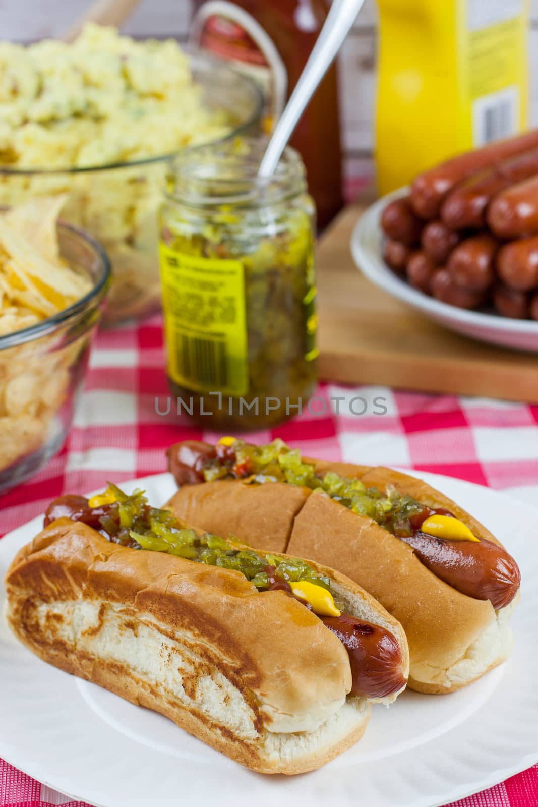 Grilled hot dogs on a paper plate sitting on a table with potato salad, chips, and condiments.