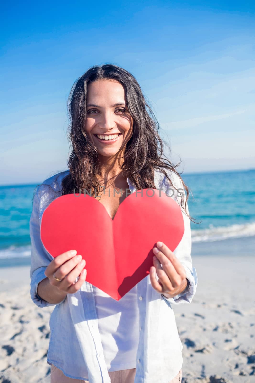 Smiling woman holding heart card at the beach by Wavebreakmedia