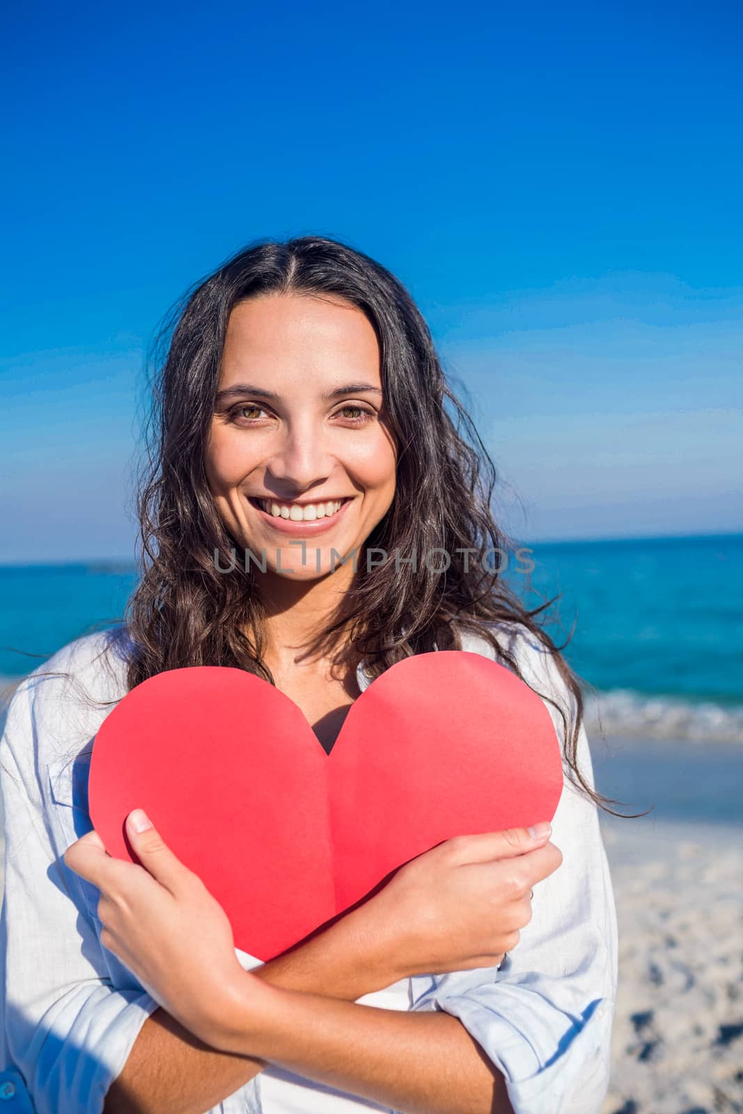 Smiling woman holding heart card at the beach on a sunny day