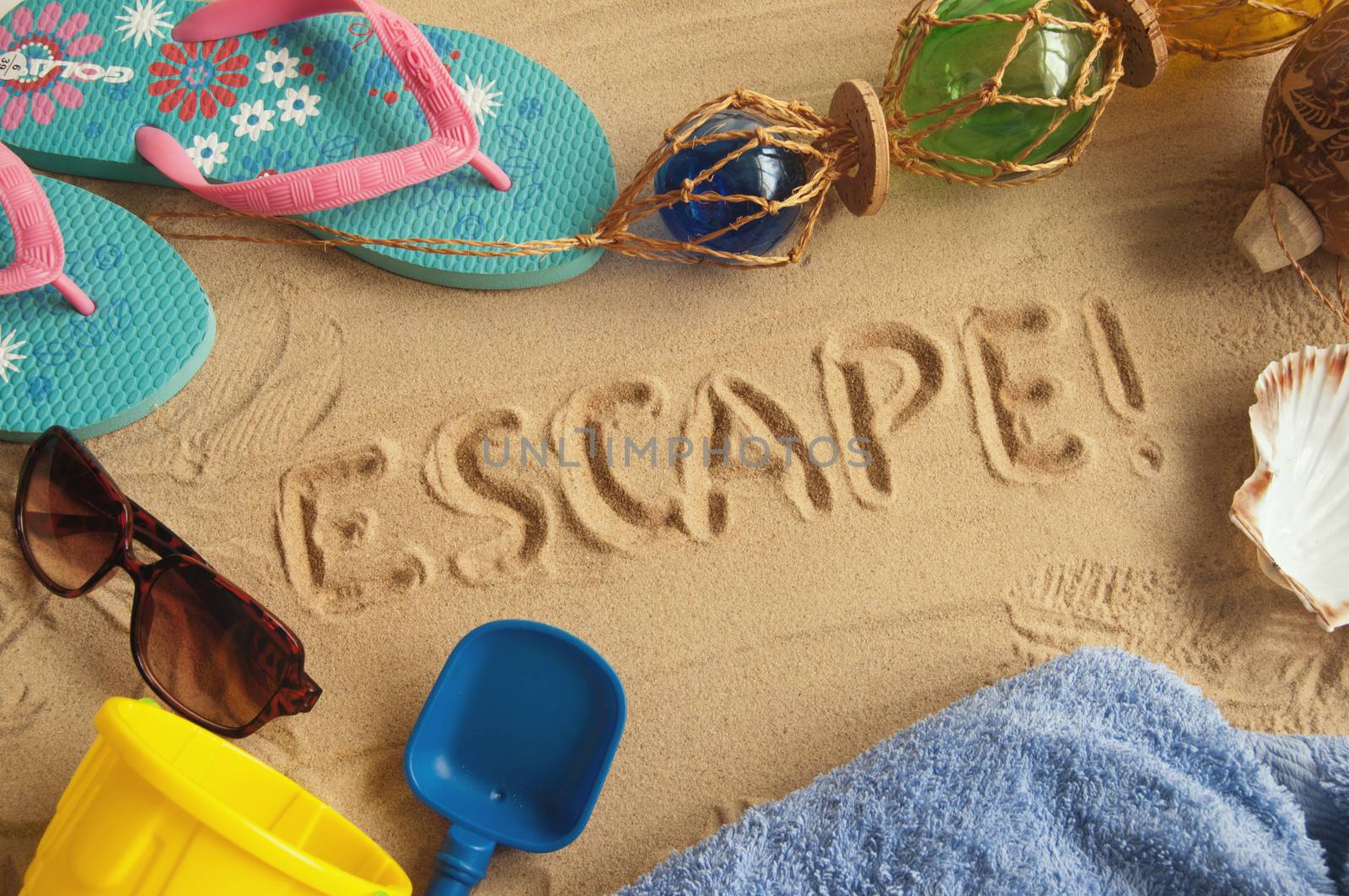 Travel concept with escape drawn in sand surrounded by beach items including, flip flops, towel, sunglasses and seashell