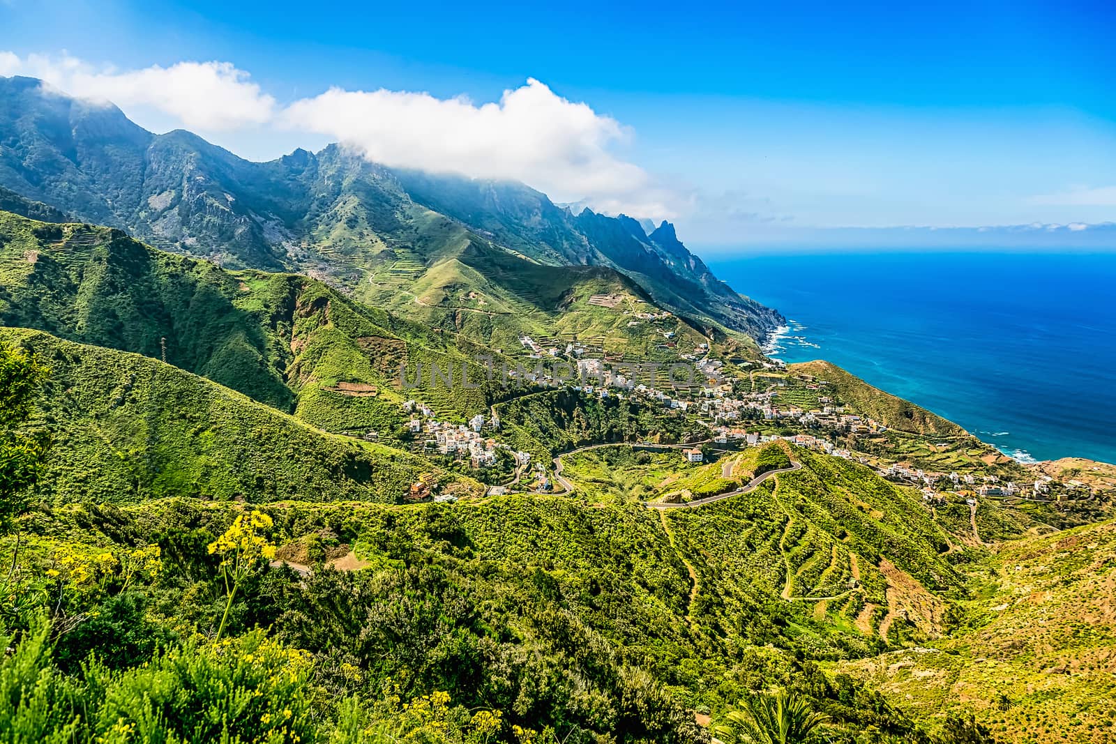 Small city or village buildings and winding or serpantine road in green mountain or rock valley and clouds with horizon landscape near coast or shore of Atlantic ocean in Tenerife Canary island, Spain