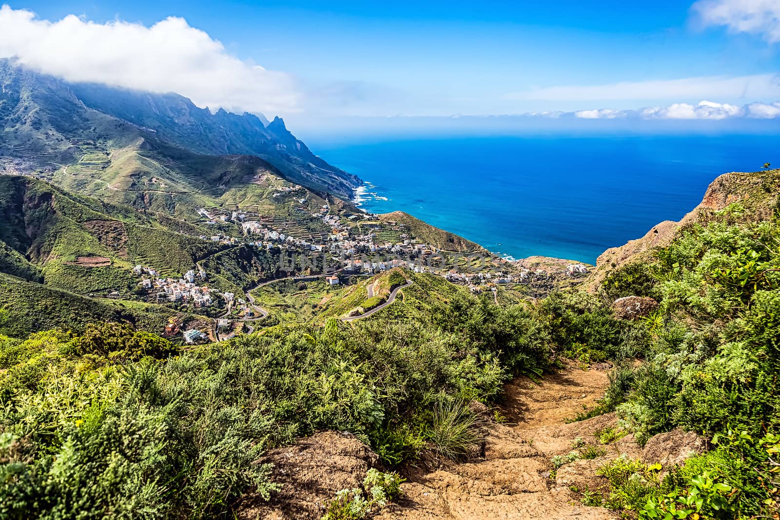 View from mountain footpath to valley with city or village buildings and winding or serpantine road and clouds with horizon landscape near coast or shore of Atlantic ocean in Tenerife Canary island, Spain