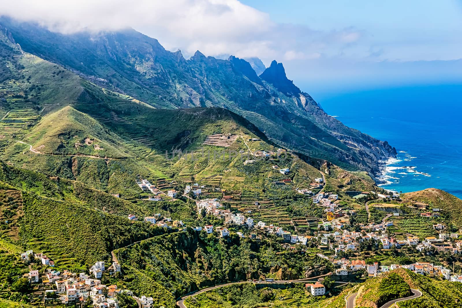 City or village buildings and winding or serpantine road in green mountain or rock valley and clouds with horizon landscape near coast of Atlantic ocean in Tenerife Canary island, Spain