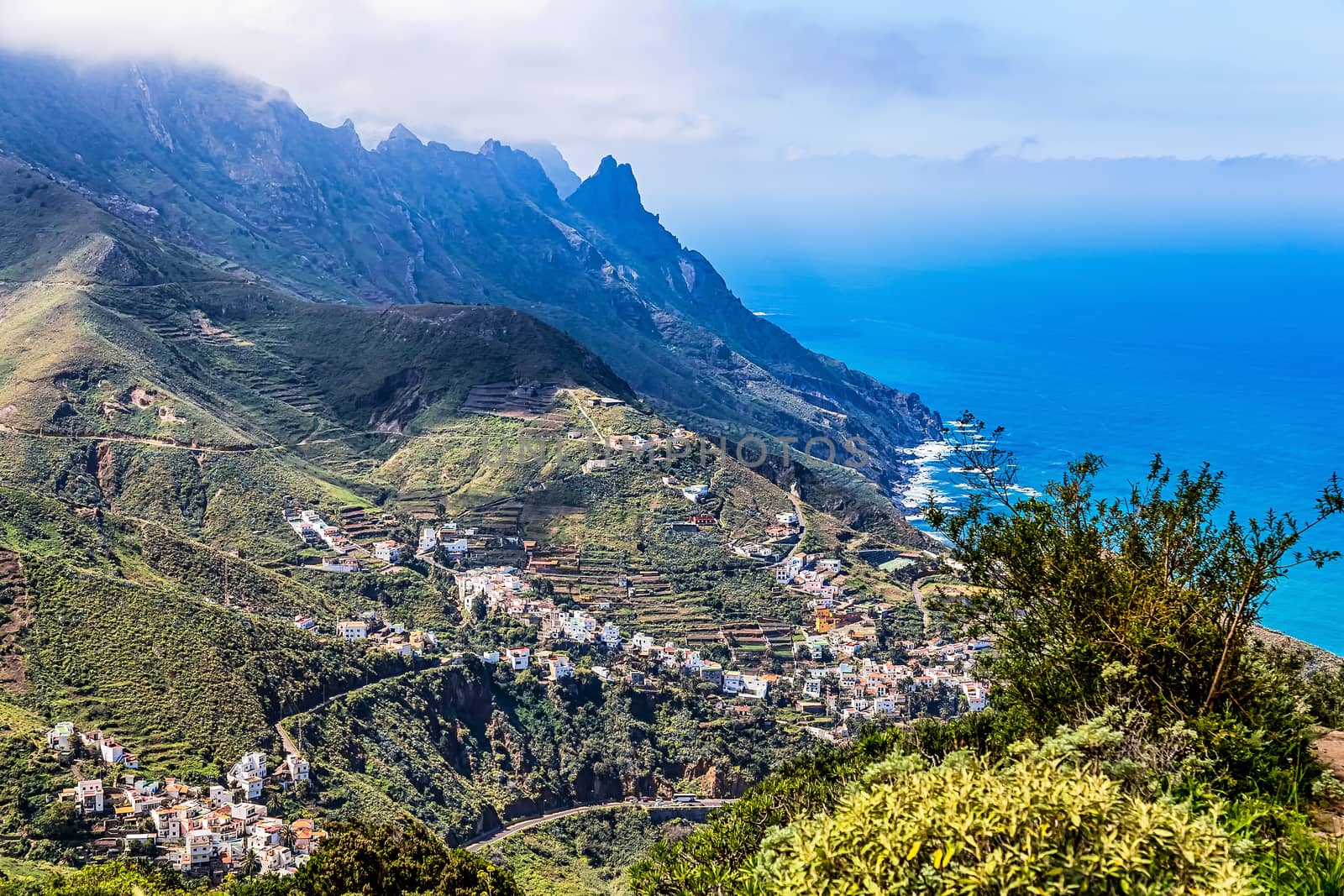 Village buildings and winding or serpantine road in green mountain or rock valley and clouds with horizon landscape near shore of Atlantic ocean in Tenerife Canary island, Spain