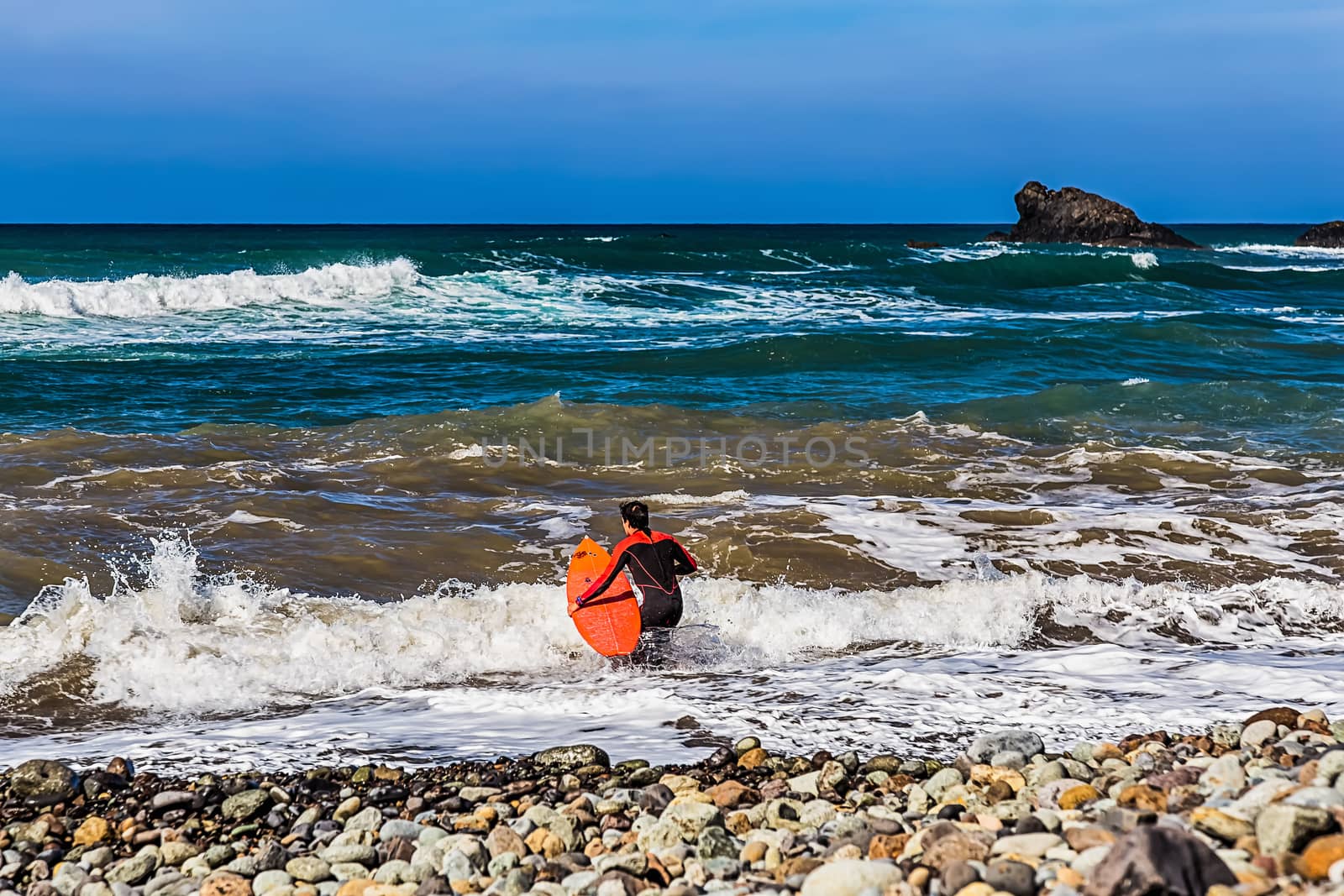 Surfer with a surfboard goes to Atlantic ocean with waves on coast or shore stone beach