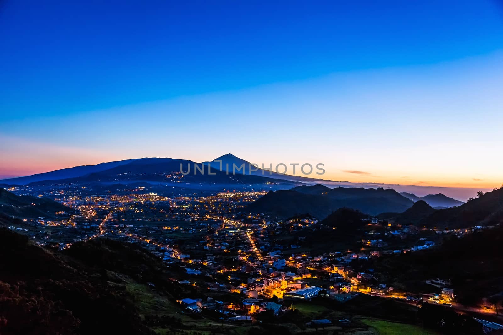 City or town with illumination after sunset or sundown at evening in mountains with blue sky and Teide volcano on background in Tenerife Canary island, Spain at spring or summer