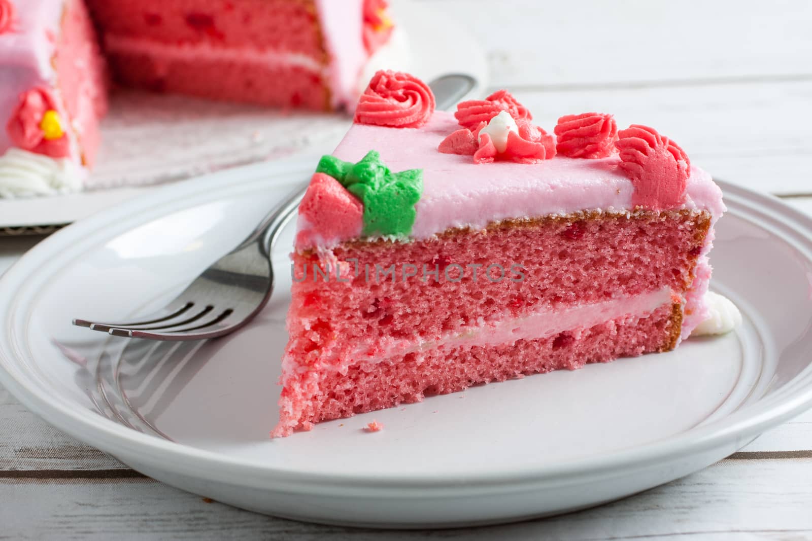 Strawberry layer cake with strawberry frosting and strawberry cream cheese filling on a white plate with a fork and a cake in the background.