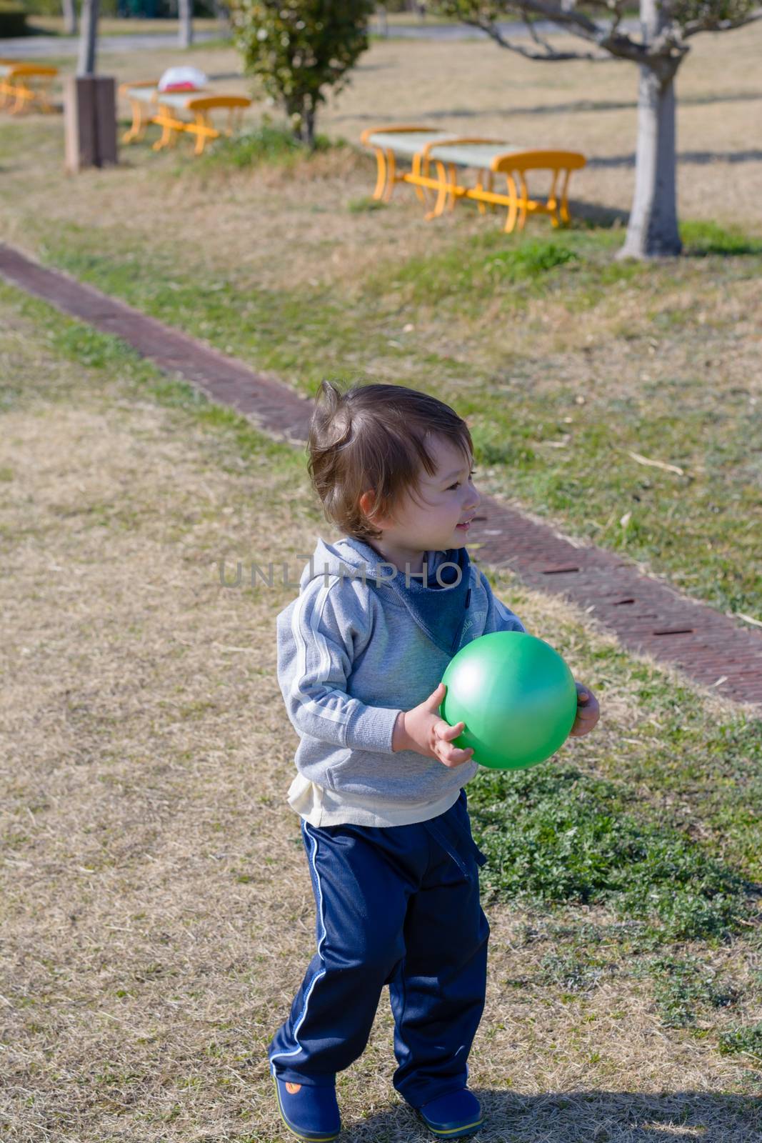 A 2 year old boy holding a ball and smiling at a playground.