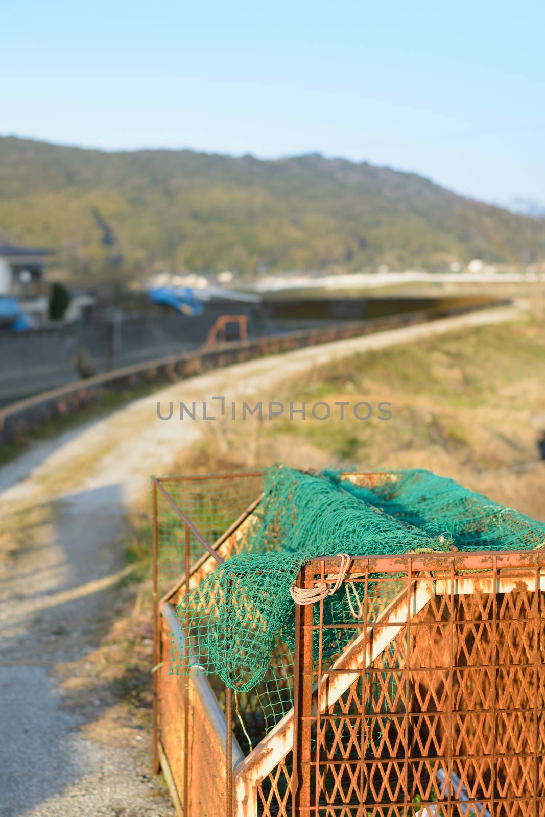 A rusty container for garbage next to a dirt path following the curve of a river with mountains in the background. In the countryside of Kochi, Japan.