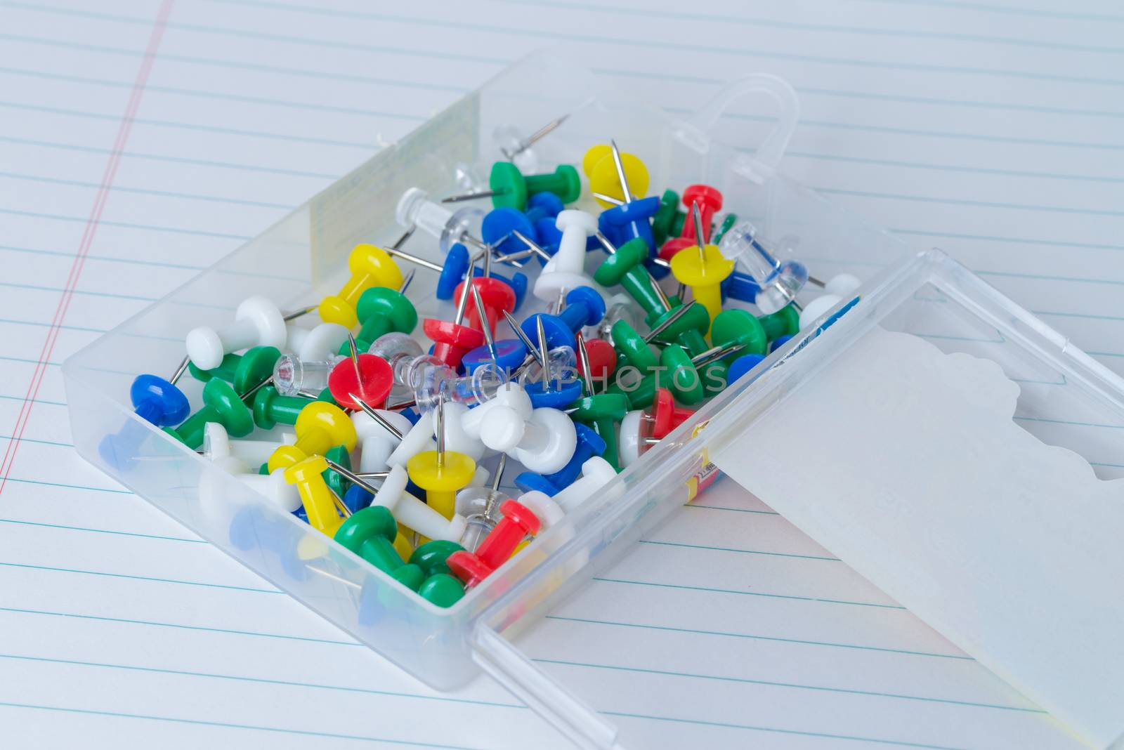 A plastic case full of multicolored thumb tacks on a white piece of lined loose leaf paper.
