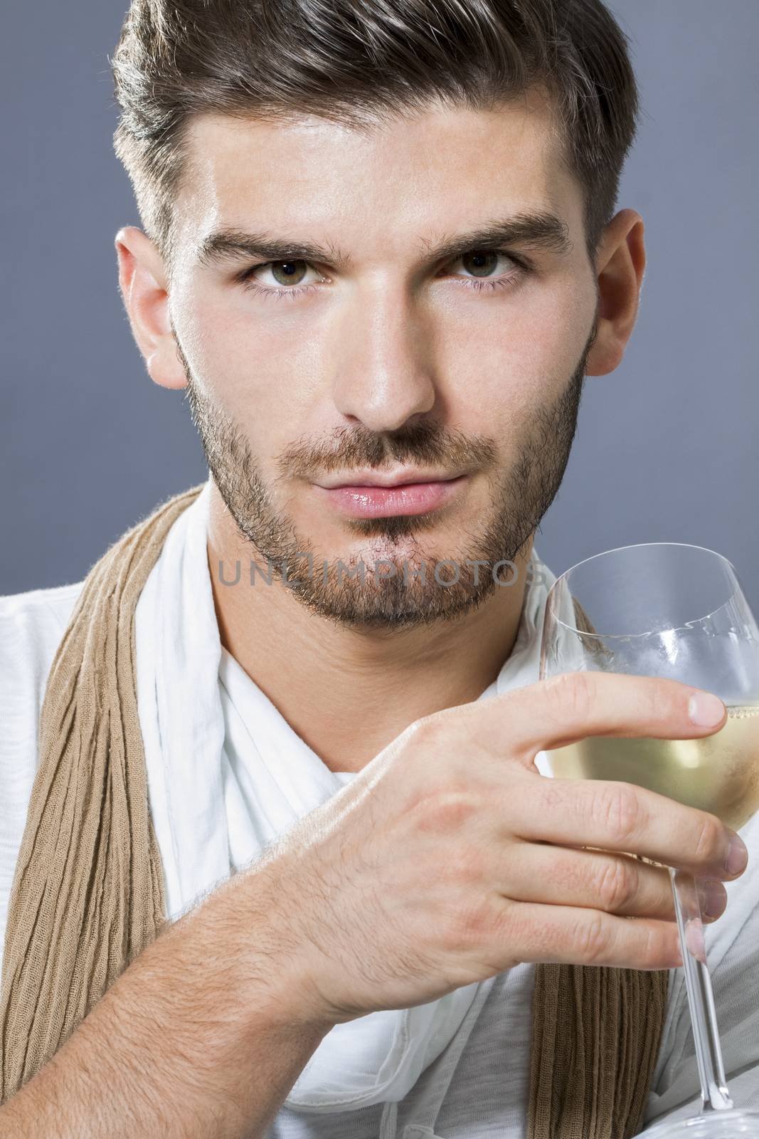 Sexy handsome young man with a beard wearing an elegant scarf drinking white wine and looking at the camera with a serious intense expression
