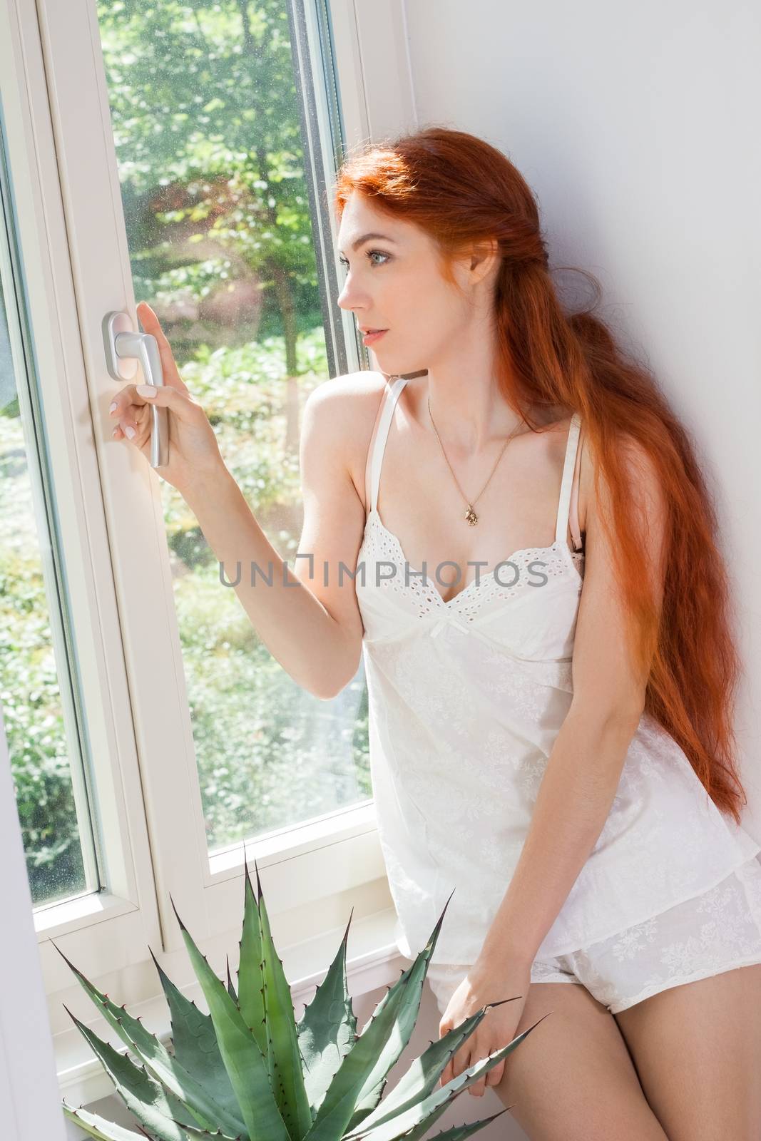 Thoughtful Pretty Young Woman with Long Blond Hair, Wearing White Night Wear, Sitting at the Glass Window While Looking Into Distance at Early in the Morning.