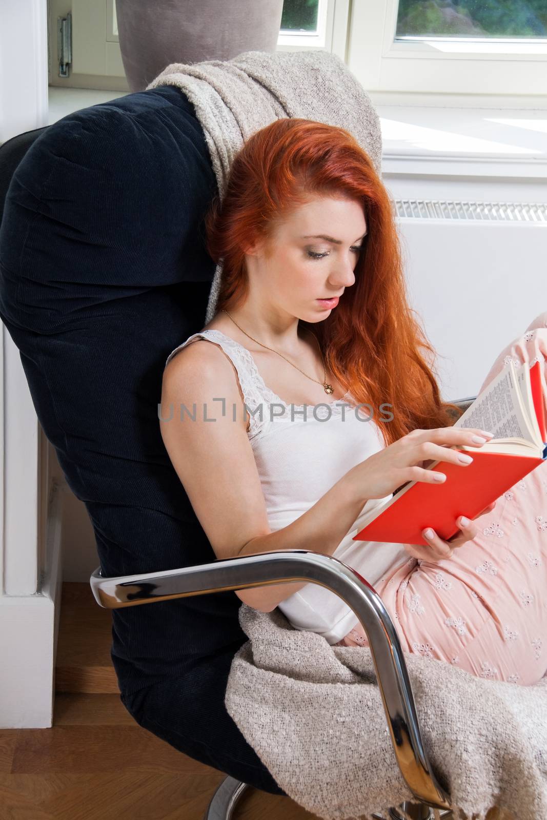Pretty Young Woman Sitting on a Chair Beside her Bed with Legs Crossed, While Reading a Red Book Seriously.