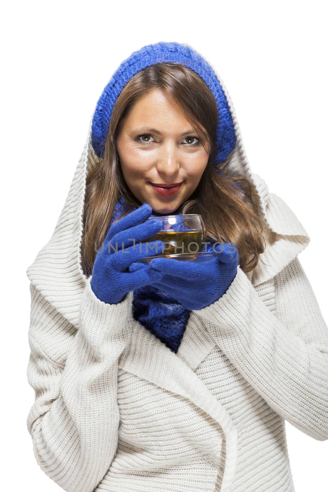Fashionable young woman in a blue knitted winter ensemble and cowl neck jersey sipping a cup of hot tea with a smile in an effort to keep warm, isolated on white