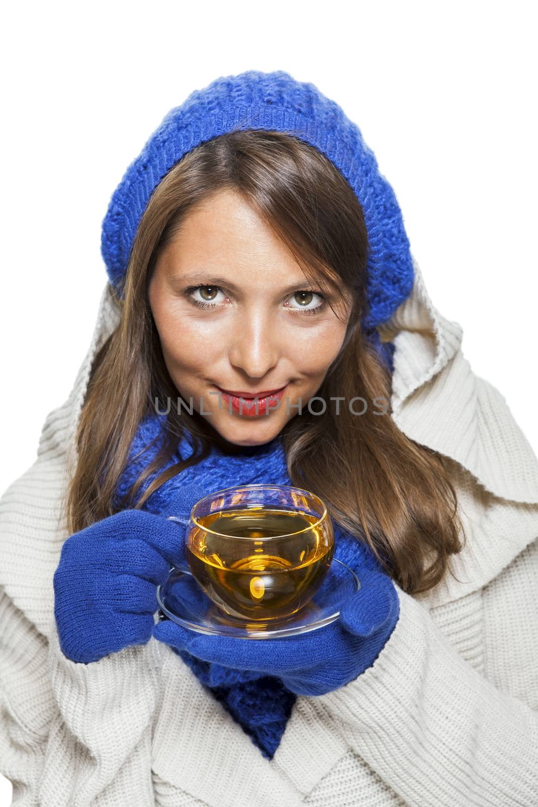 Fashionable young woman in a blue knitted winter ensemble and cowl neck jersey sipping a cup of hot tea with a smile in an effort to keep warm, isolated on white