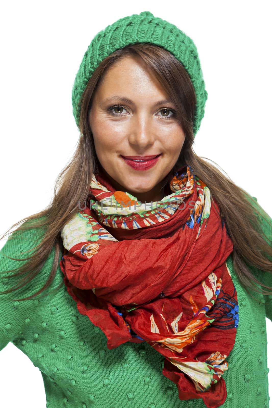 Cute sexy young woman with long brunette hair in a green winter outfit smiling playfully at the camera with her hand to her red scarf, on white