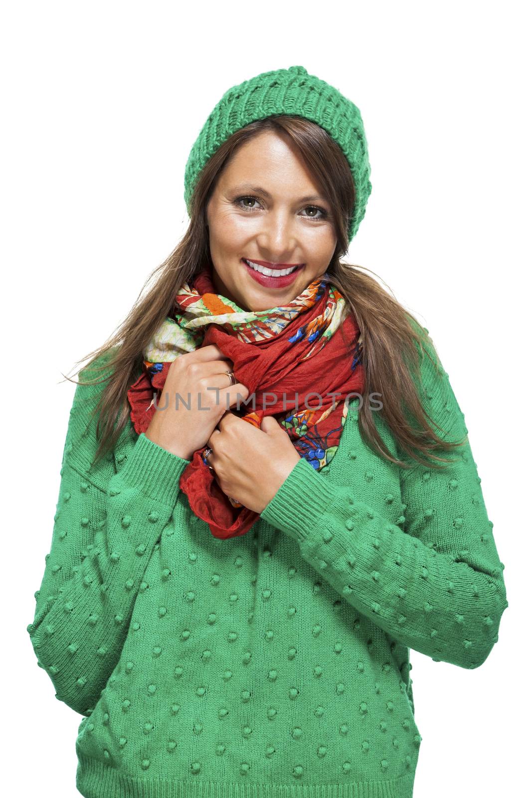 Cute sexy young woman with long brunette hair in a green winter outfit smiling playfully at the camera with her hand to her red scarf, on white