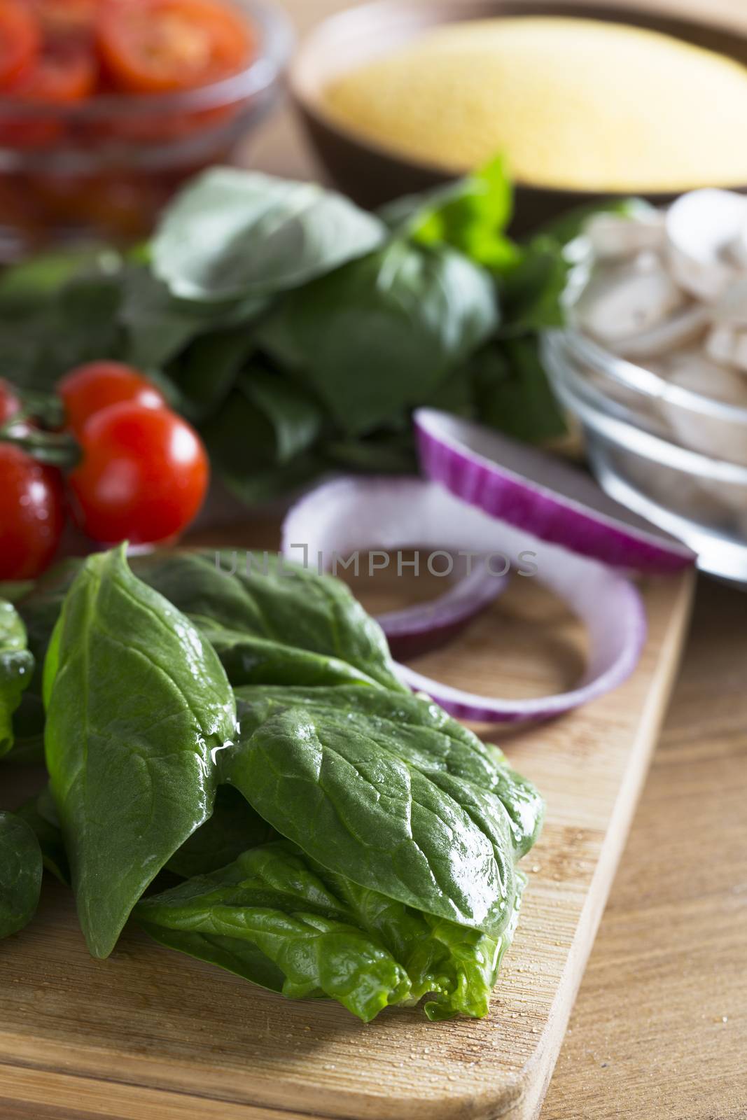 Fresh ingredients for making polenta dish.  Spinach in the foreground with red onion slices, cherry tomatoes, mushrooms and polenta in the background  out of focus.  Vertical image.