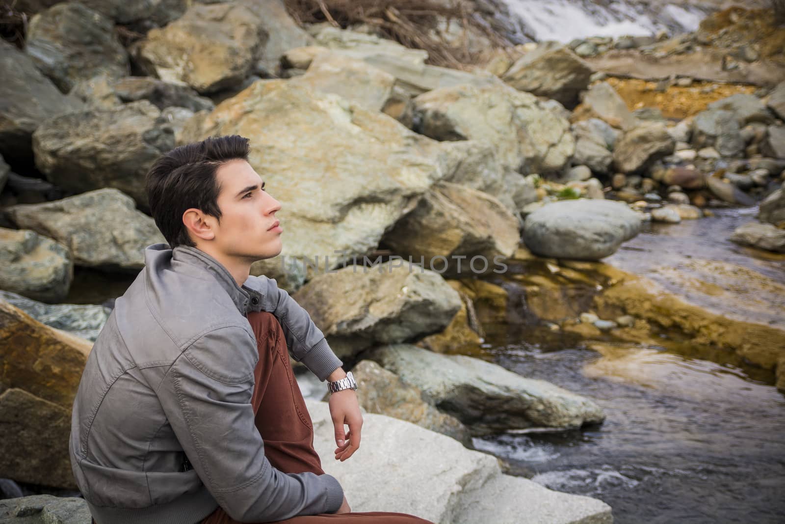 Handsome young man outdoor sitting alone at river or water stream looking up thinking