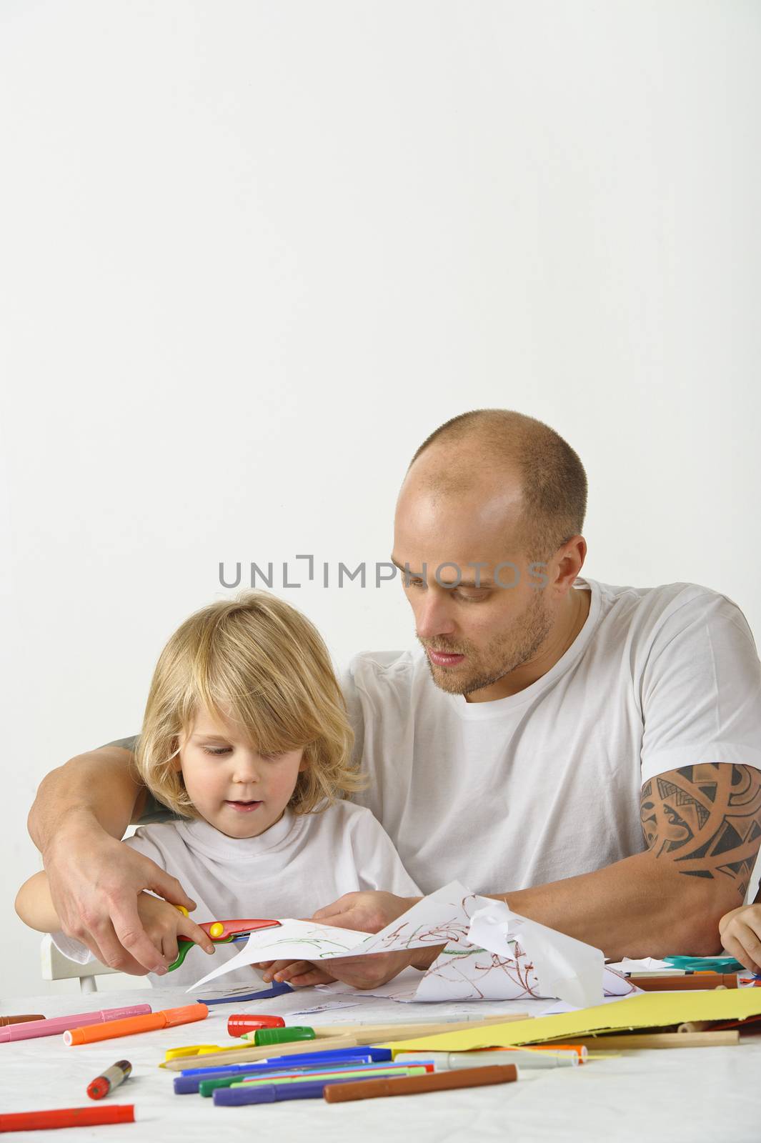 Father teaches his young son how to use a scissors. The father's upper arms are tattoed.