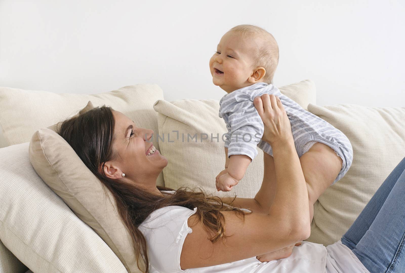 Mother lifts her baby son in the air. She's lying on a sofa. They're laughing
