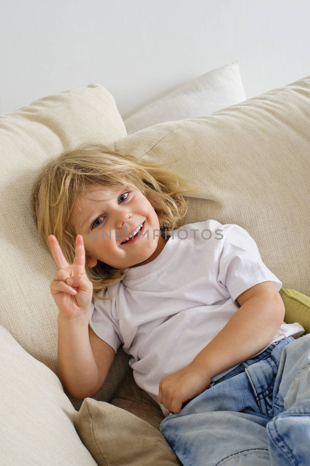 Young boy sitting on sofa looking at camera. He's giving the V sign with his fingers, palm towards the camera
