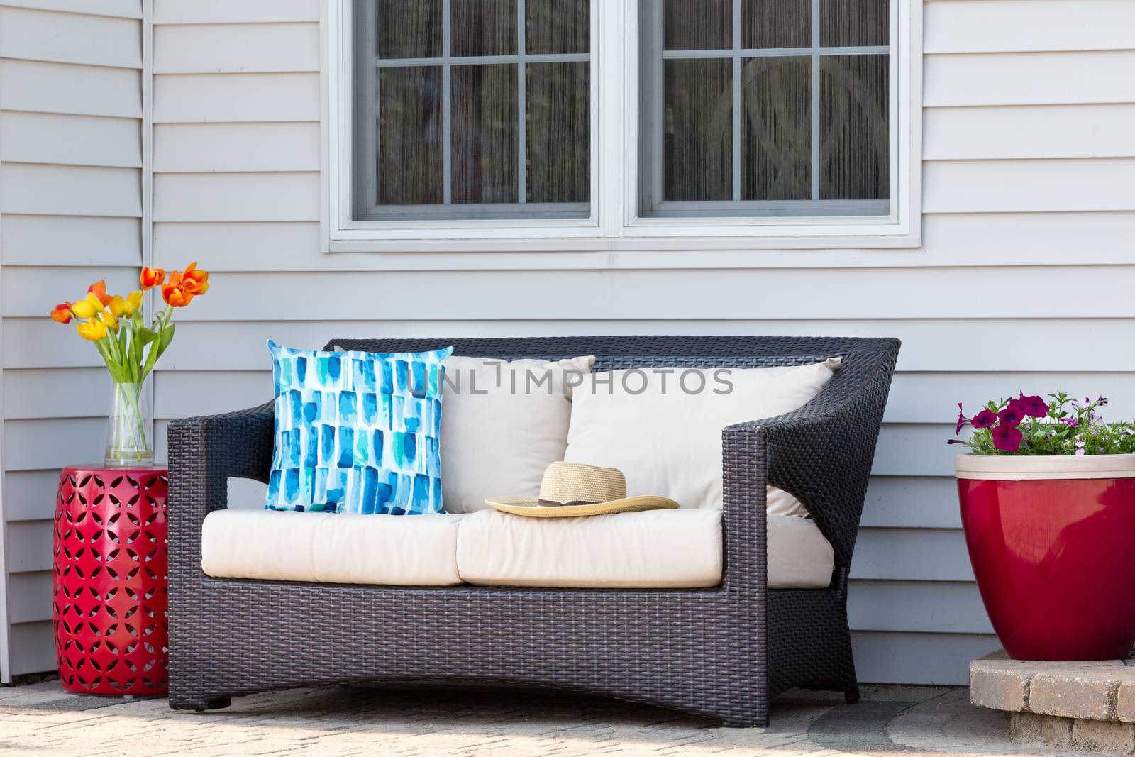 Comfortable outdoor living area on a brick patio with a deep seating settee and cushions flanked by red ceramic pedestal table and flowerpot with spring flowers and a straw sunhat