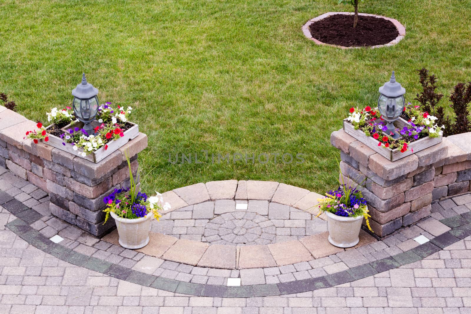 Curved entrance to an outdoors patio with shallow steps leading to a brick paved living area flanked by two pillars with lamps and colorful flowers in square pots, high angle view with green grass