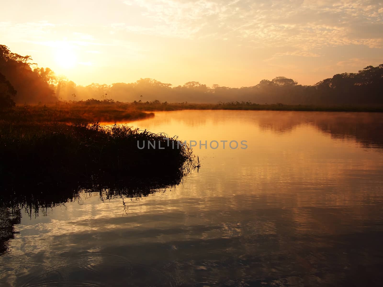 An orange sunrise with some light fog over the water of the Tambopata river during sunrise in the Amazon rainforest in eastern Peru near the border with Brazil.